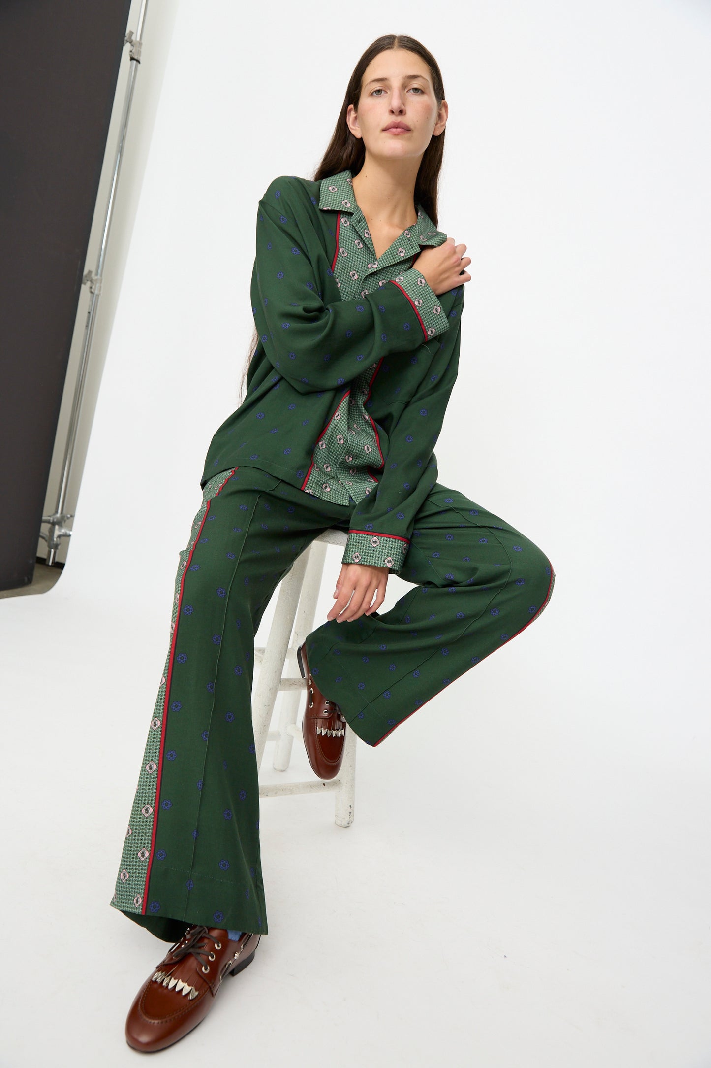 A person wearing the TOGA ARCHIVES Inner Print Drawstring Pant in Green sits on a white stool against a white studio background, paired with brown shoes.