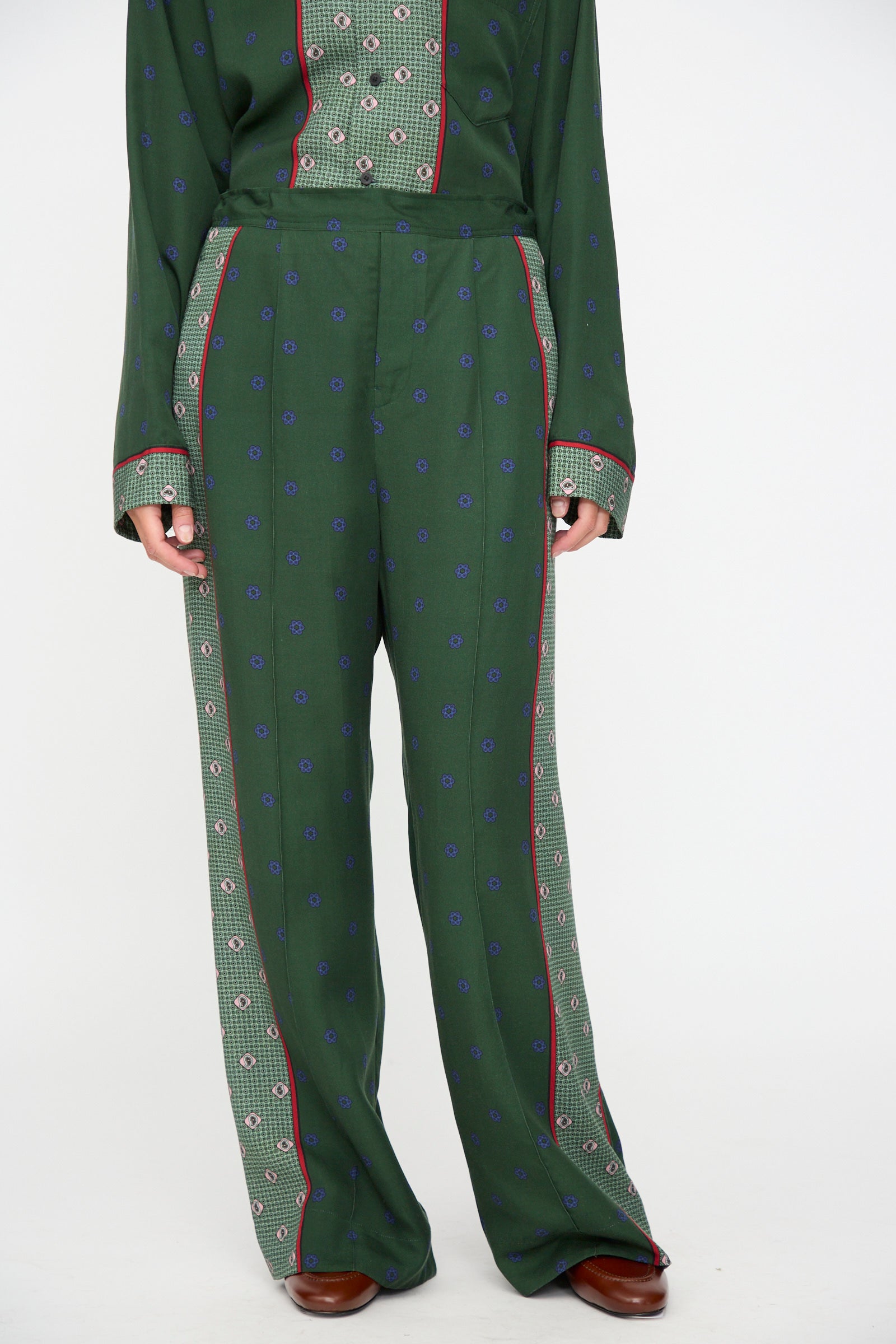 A person is wearing the TOGA ARCHIVES Inner Print Drawstring Pant in Green, featuring a green floral pattern with red and blue accents, paired with a green patterned long-sleeve shirt and brown shoes, photographed against a white background.