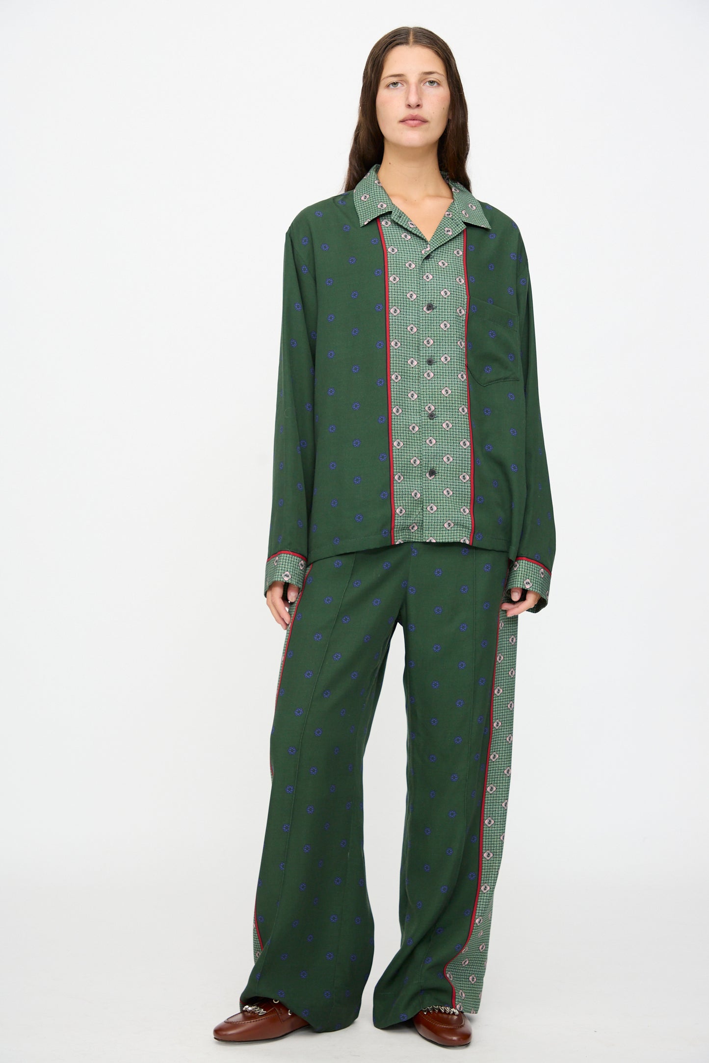 A person stands against a plain white background wearing the TOGA ARCHIVES Inner Print Long Sleeve Shirt in Green, paired with pajama-style bottoms featuring mismatched geometric prints and brown shoes.