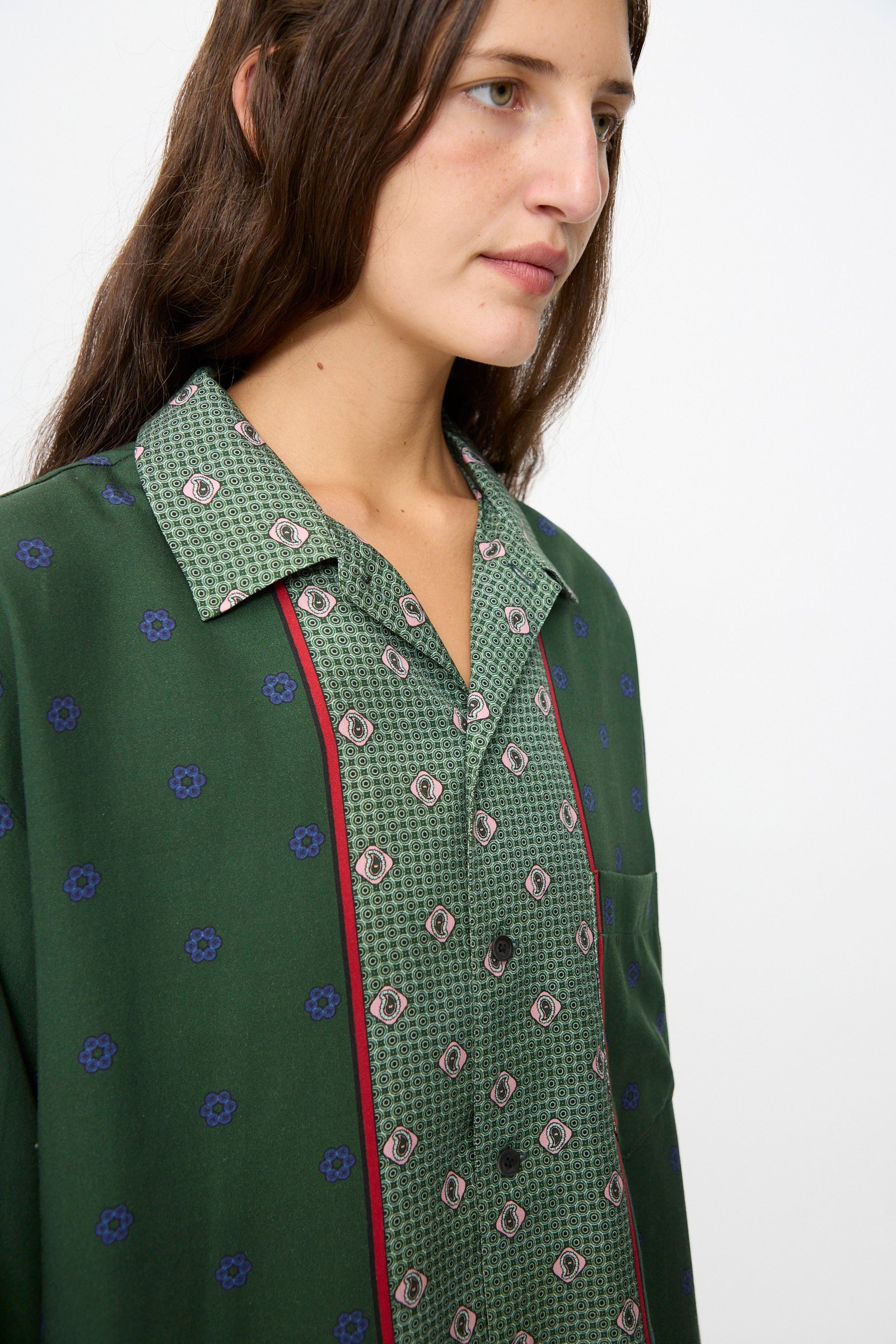 A person with long brown hair wears the TOGA ARCHIVES Inner Print Long Sleeve Shirt in Green, featuring blue and red accents, while looking away from the camera against a plain background.
