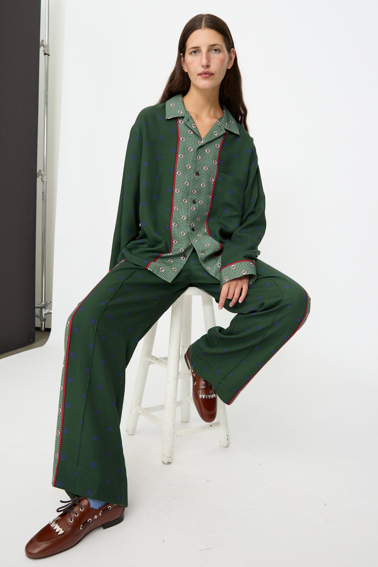 A person wearing the TOGA ARCHIVES Inner Print Long Sleeve Shirt in Green and brown shoes, sitting on a white stool against a plain white background.
