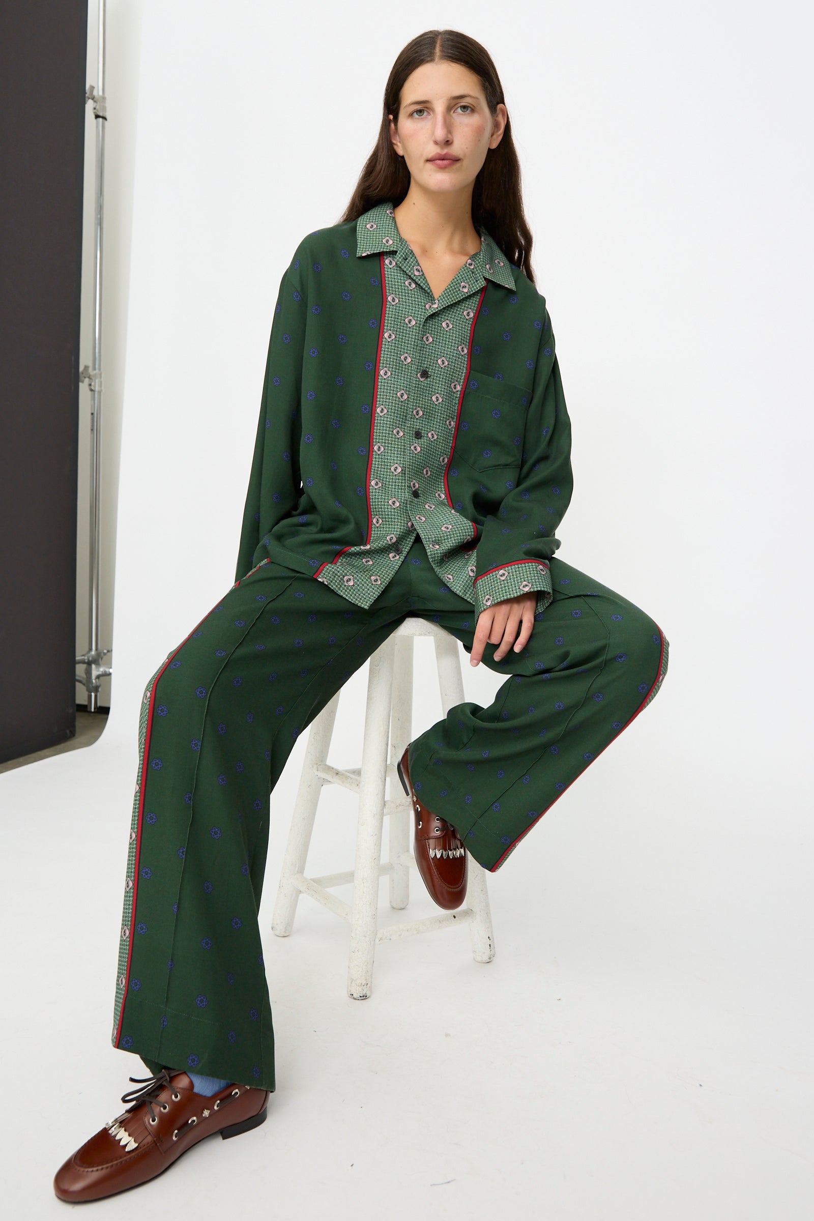 A person wearing the TOGA ARCHIVES Inner Print Long Sleeve Shirt in Green and brown shoes, sitting on a white stool against a plain white background.