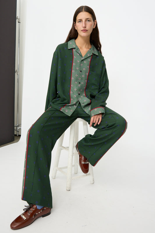 A person wearing the TOGA ARCHIVES Inner Print Long Sleeve Shirt in Green and brown shoes, sitting on a white stool against a plain white background.