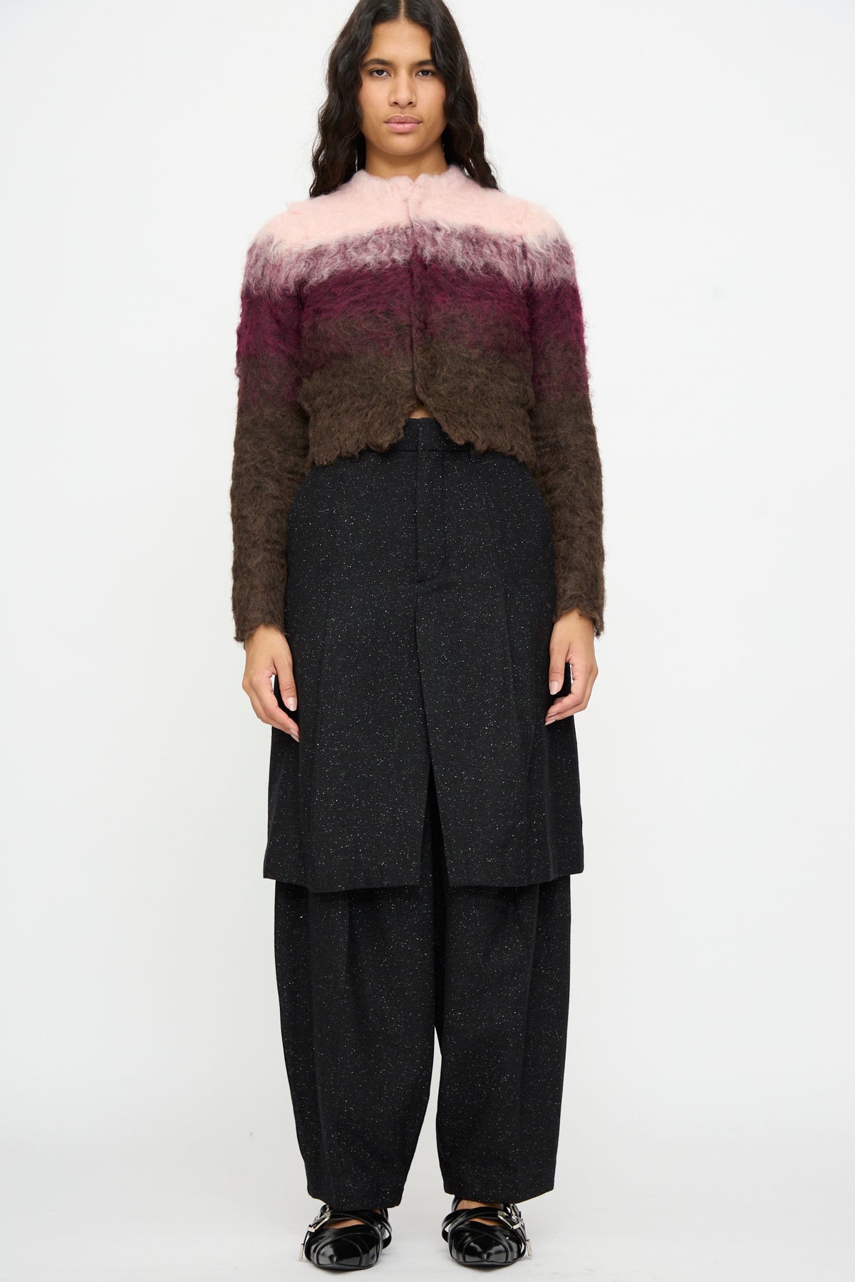 A person stands facing forward wearing a fuzzy pink and brown speckled wool sweater, paired with TOGA ARCHIVES Layered Trousers in Black and black shiny shoes, against a plain white background.