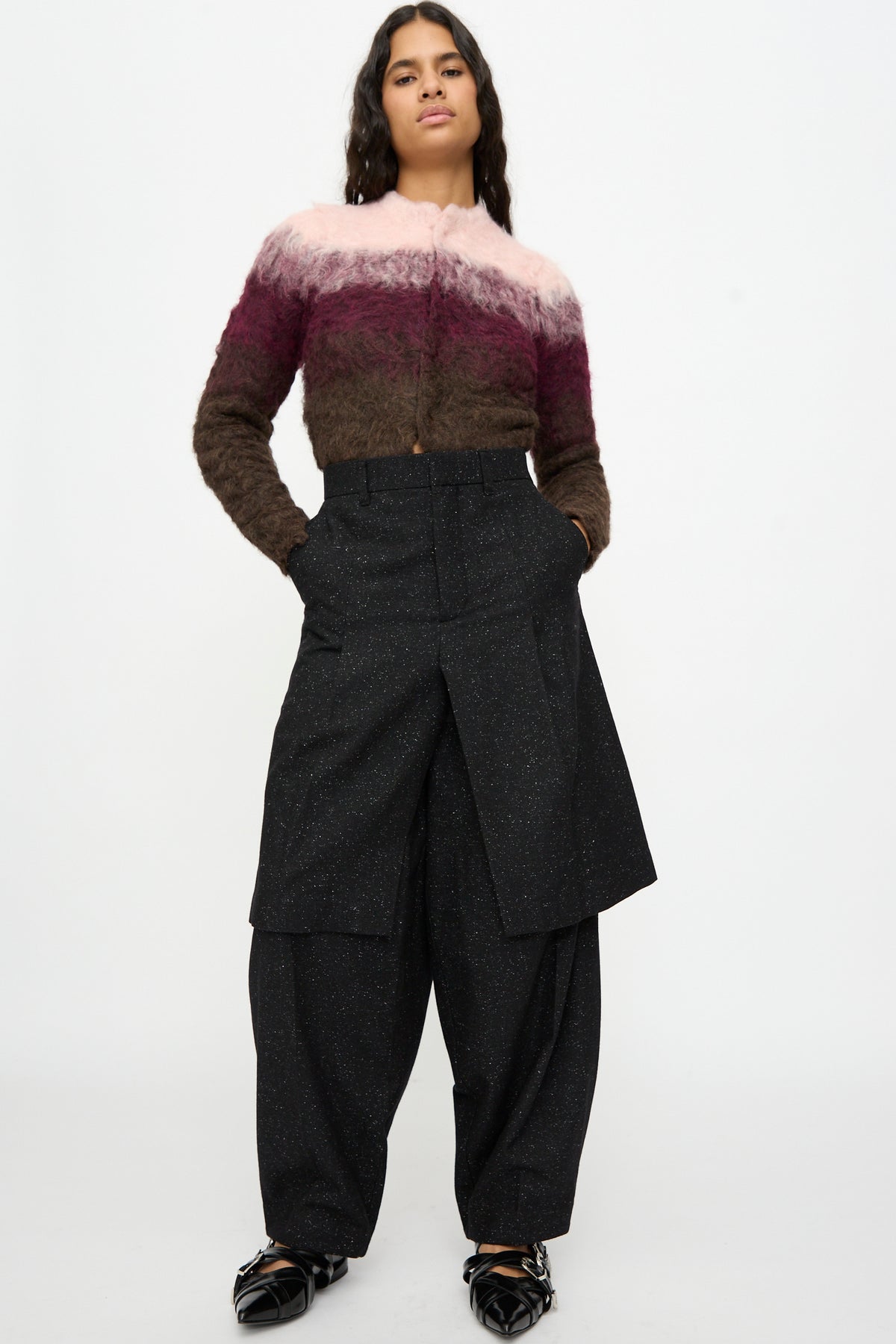 A person stands against a plain background wearing a fuzzy pink and brown speckled wool sweater, TOGA ARCHIVES Layered Trousers in Black, and black shoes.