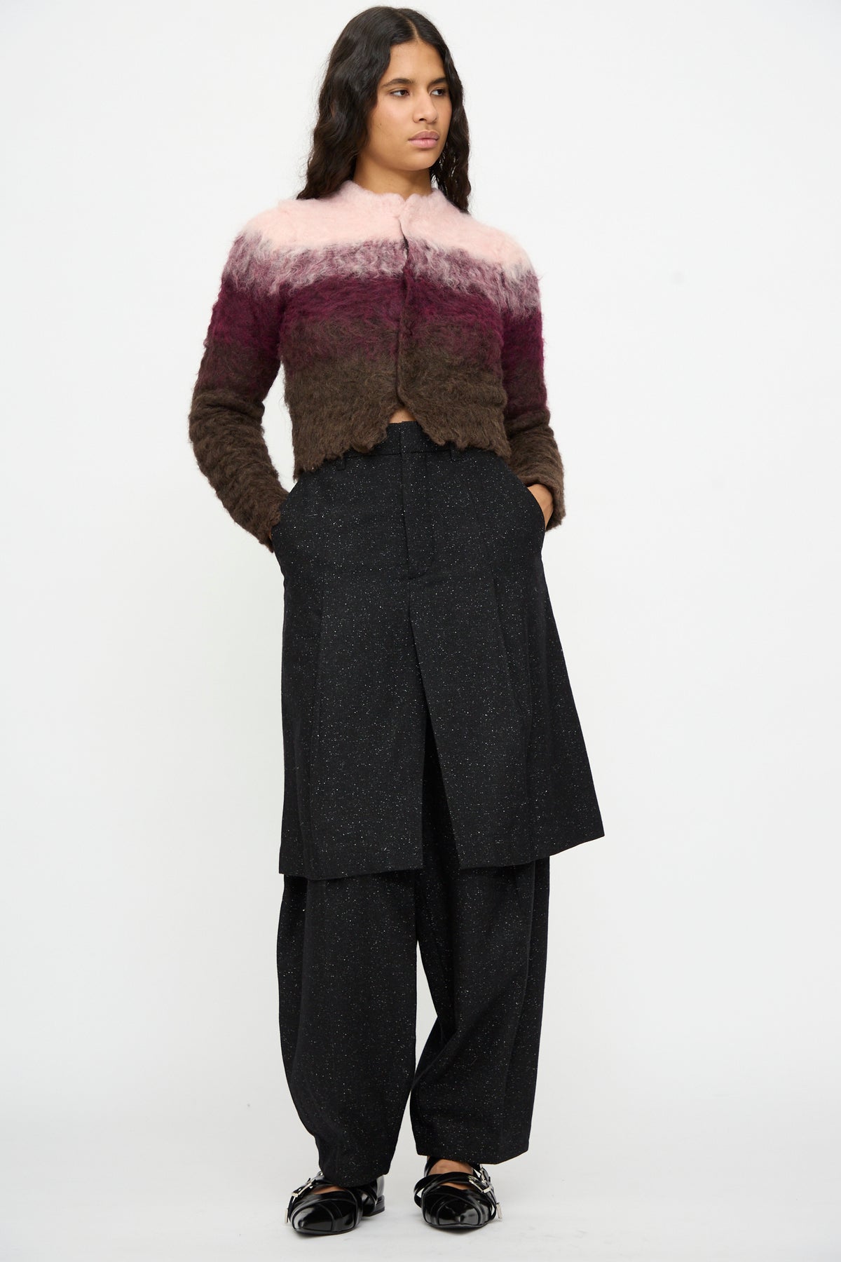 A model wears a fuzzy, color-blocked cardigan in shades of pink and brown, paired with TOGA ARCHIVES’ Layered Trousers in Black and black shoes, standing with hands in pockets against a plain background.