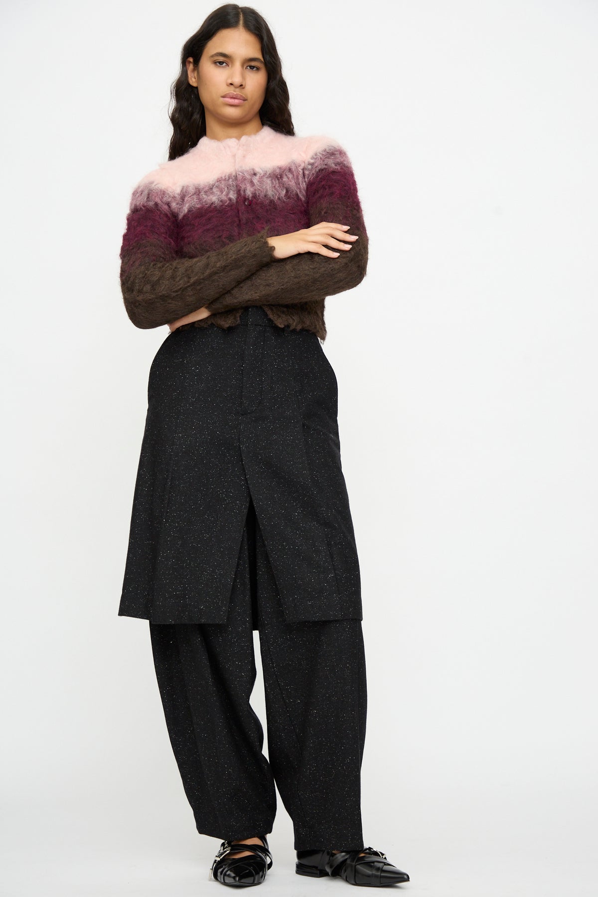 A person stands against a plain background wearing a fuzzy, ombre sweater in pink and burgundy, TOGA ARCHIVES Layered Trousers in Black with long speckled wool fabric and a unique trouser overlay, and black shoes. This distinctive outfit is expertly made in Japan.