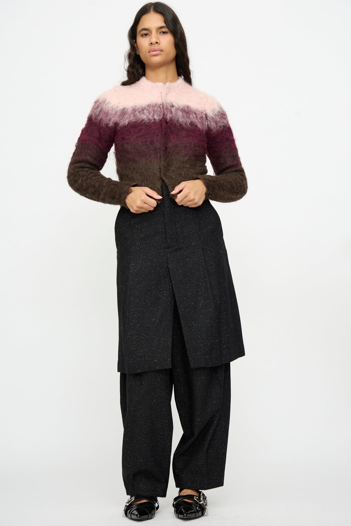 A person stands against a plain background wearing a fuzzy pink, burgundy, and brown sweater, paired with TOGA ARCHIVES Layered Trousers in Black—crafted from speckled wool—and black shoes.