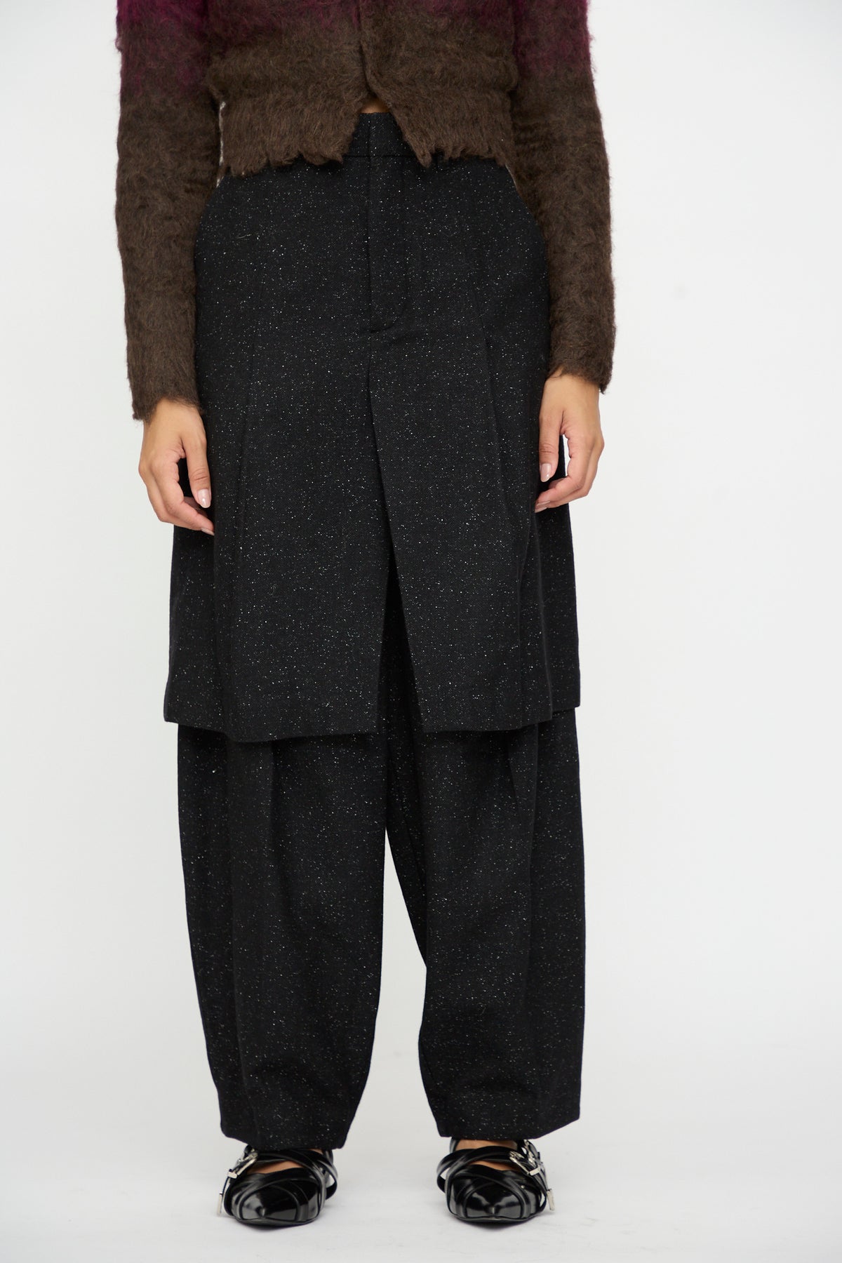 A person wearing TOGA ARCHIVES Layered Trousers in Black, styled with a brown fuzzy speckled wool sweater and black shoes, stands against a white background.