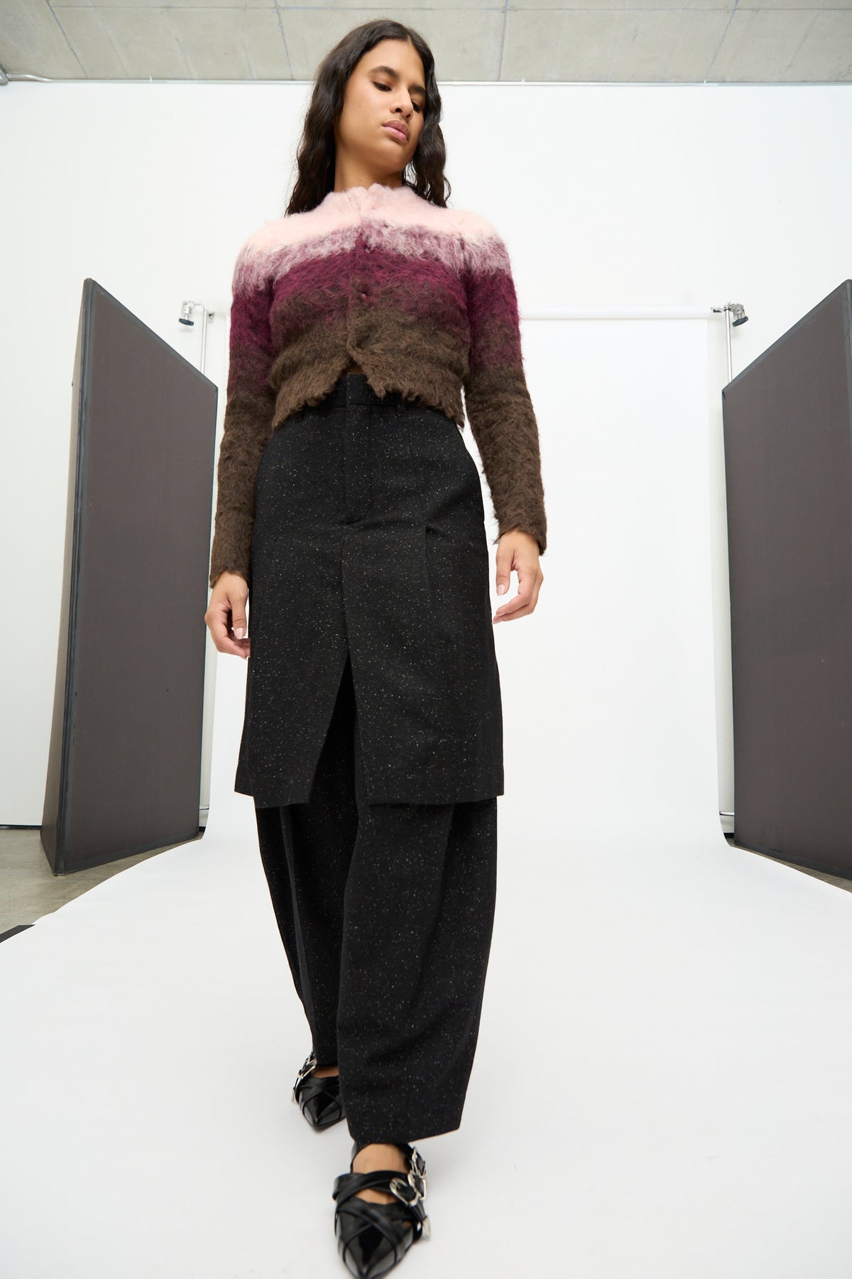 A woman wearing a fuzzy, speckled wool color-block sweater and wide-leg black pants stands on a white runway between two black panels, looking slightly downward. Her look is elevated with the Layered Trousers in Black from TOGA ARCHIVES, evoking understated sophistication.