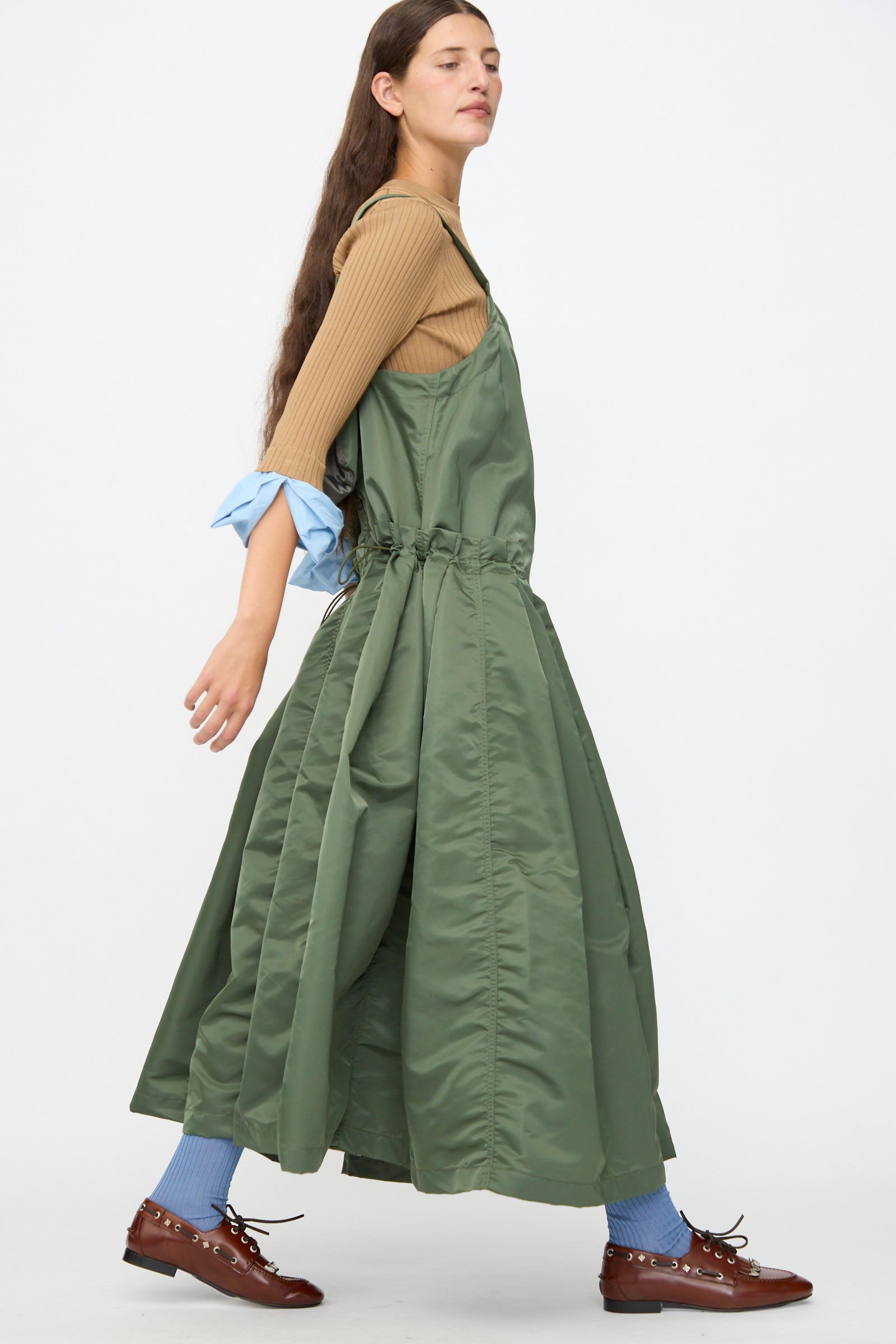 A woman is shown in profile wearing the TOGA ARCHIVES Nylon Twill Dress in Khaki—a long, A-line silhouette maxi dress styled over a tan top, with blue socks, brown shoes, and featuring a blue sleeve detail; the background is plain white.
