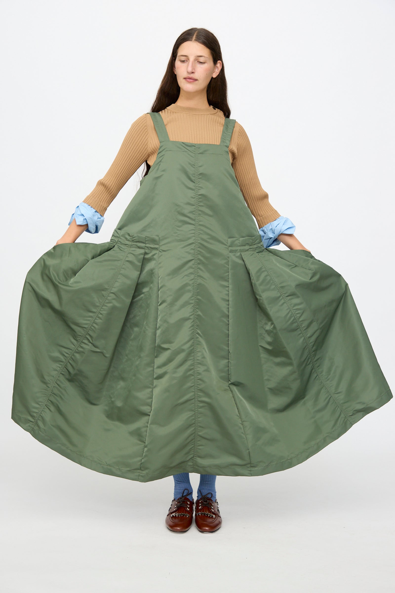 A woman stands holding out the sides of the TOGA ARCHIVES Nylon Twill Dress in Khaki, which features an A-line silhouette and maxi length, styled over a tan ribbed sweater with blue socks and brown shoes against a plain light background.
