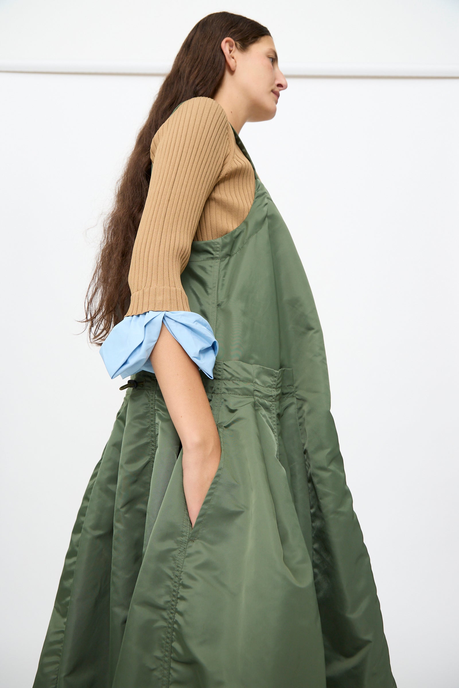 A woman stands sideways wearing the TOGA ARCHIVES Nylon Twill Dress in Khaki, featuring an elegant A-line silhouette and a voluminous maxi length, her hand tucked casually into the pocket.