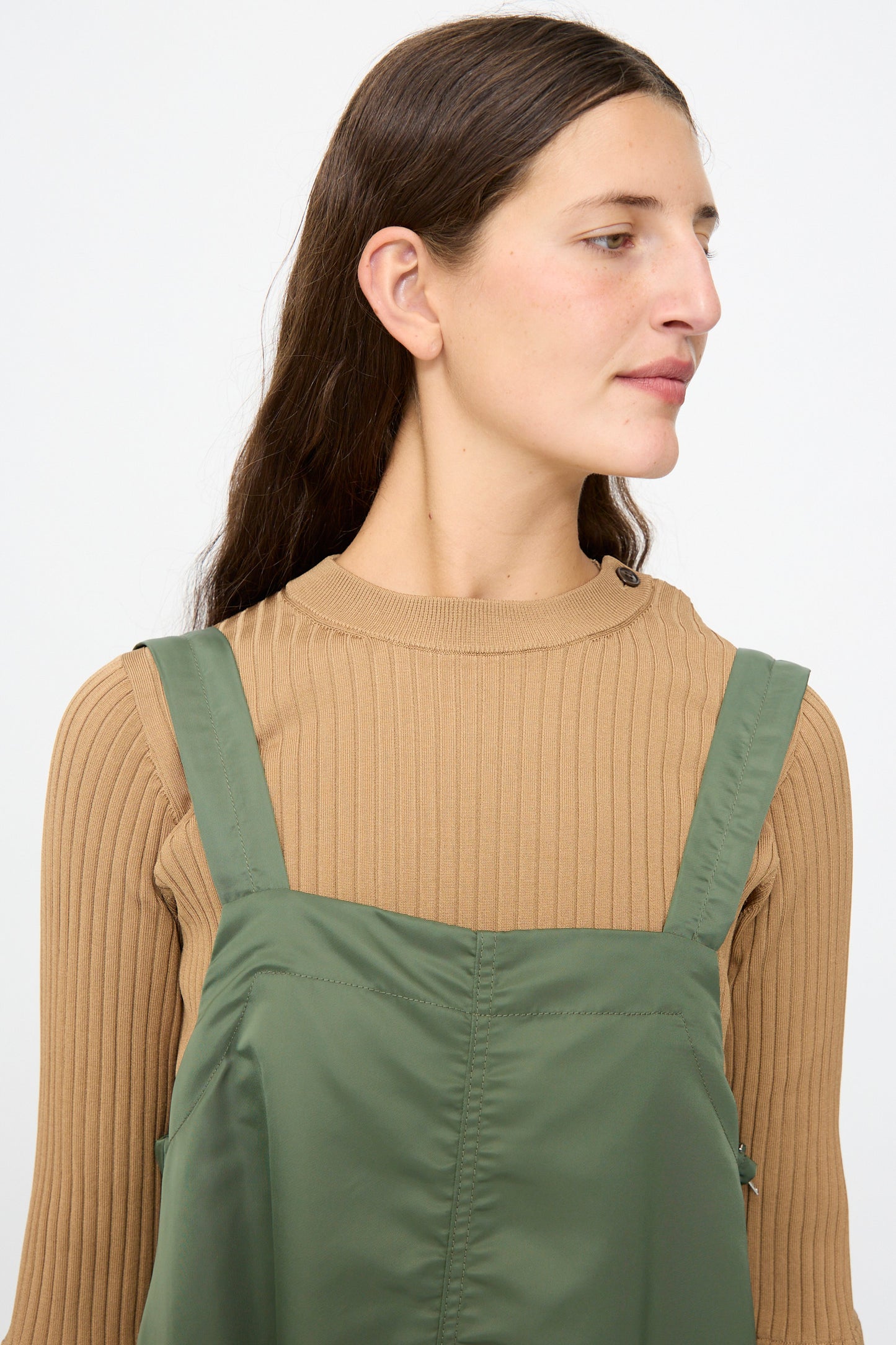 A person with long brown hair wears a beige ribbed sweater layered under the TOGA ARCHIVES Nylon Twill Dress in Khaki, which features an A-line silhouette, while looking to the side against a plain light background.
