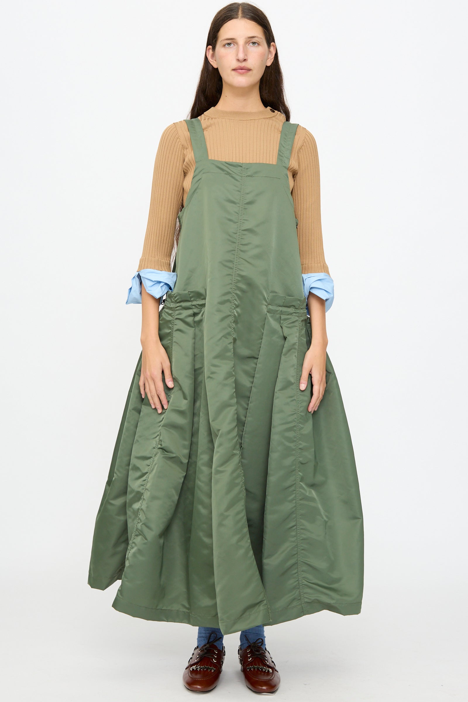 A person stands facing forward against a plain white background, wearing the TOGA ARCHIVES Nylon Twill Dress in Khaki—a pinafore-style maxi dress with an A-line silhouette. The dress is layered over a tan long-sleeve shirt with light blue cuffs and paired with blue socks and brown shoes.