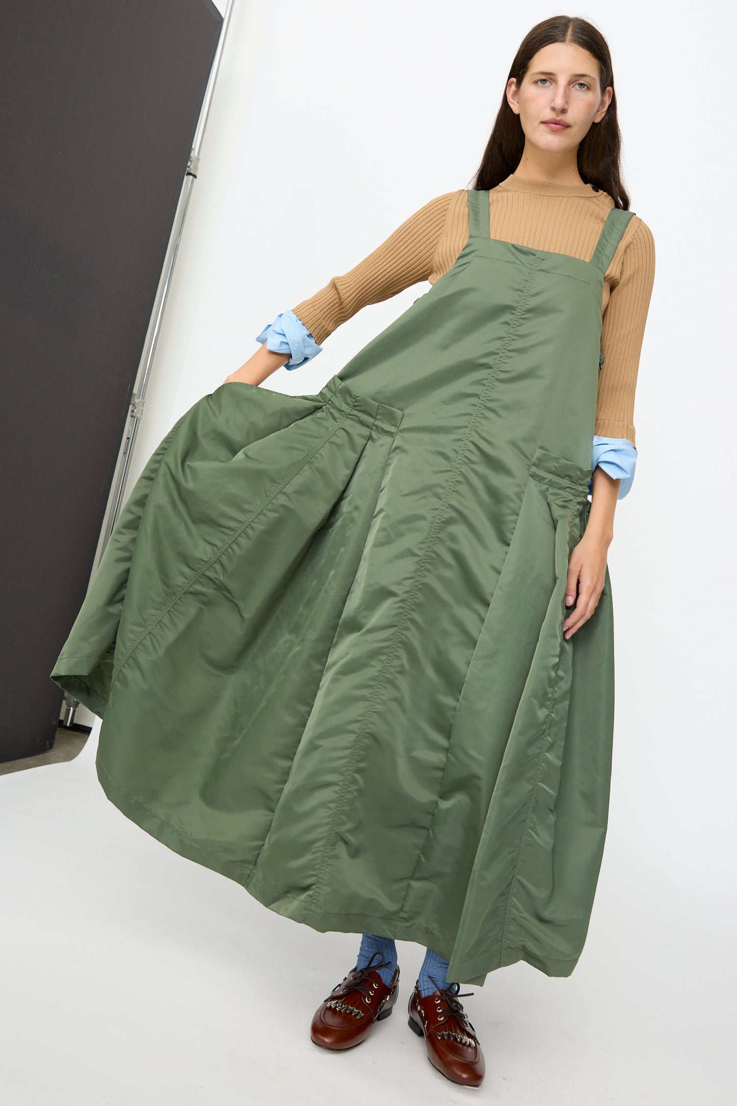 A woman stands indoors wearing the TOGA ARCHIVES Nylon Twill Dress in Khaki—a long, loose A-line silhouette—styled over a brown top and blue pants, paired with brown shoes and a neutral expression.