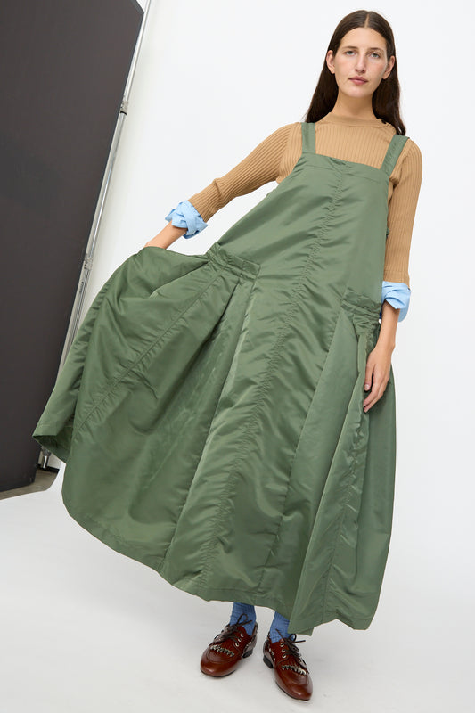 A woman stands indoors wearing the TOGA ARCHIVES Nylon Twill Dress in Khaki—a long, loose A-line silhouette—styled over a brown top and blue pants, paired with brown shoes and a neutral expression.