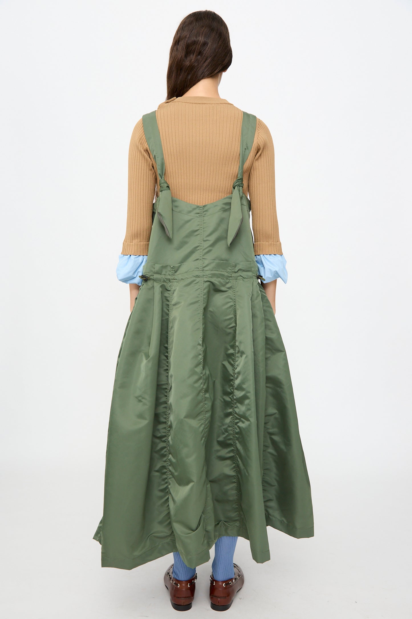 A person stands with their back to the camera against a plain white background, wearing the TOGA ARCHIVES Nylon Twill Dress in Khaki—a green sleeveless maxi dress featuring an elegant A-line silhouette and long, flared skirt—layered over a tan ribbed top and paired with brown shoes.