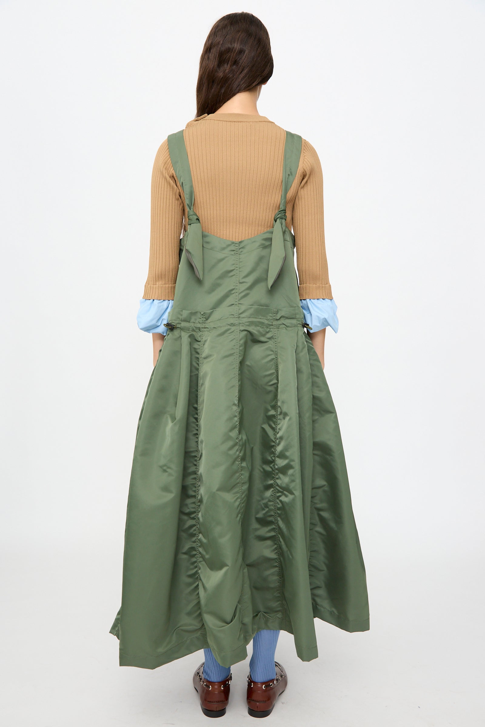 A person stands with their back to the camera against a plain white background, wearing the TOGA ARCHIVES Nylon Twill Dress in Khaki—a green sleeveless maxi dress featuring an elegant A-line silhouette and long, flared skirt—layered over a tan ribbed top and paired with brown shoes.