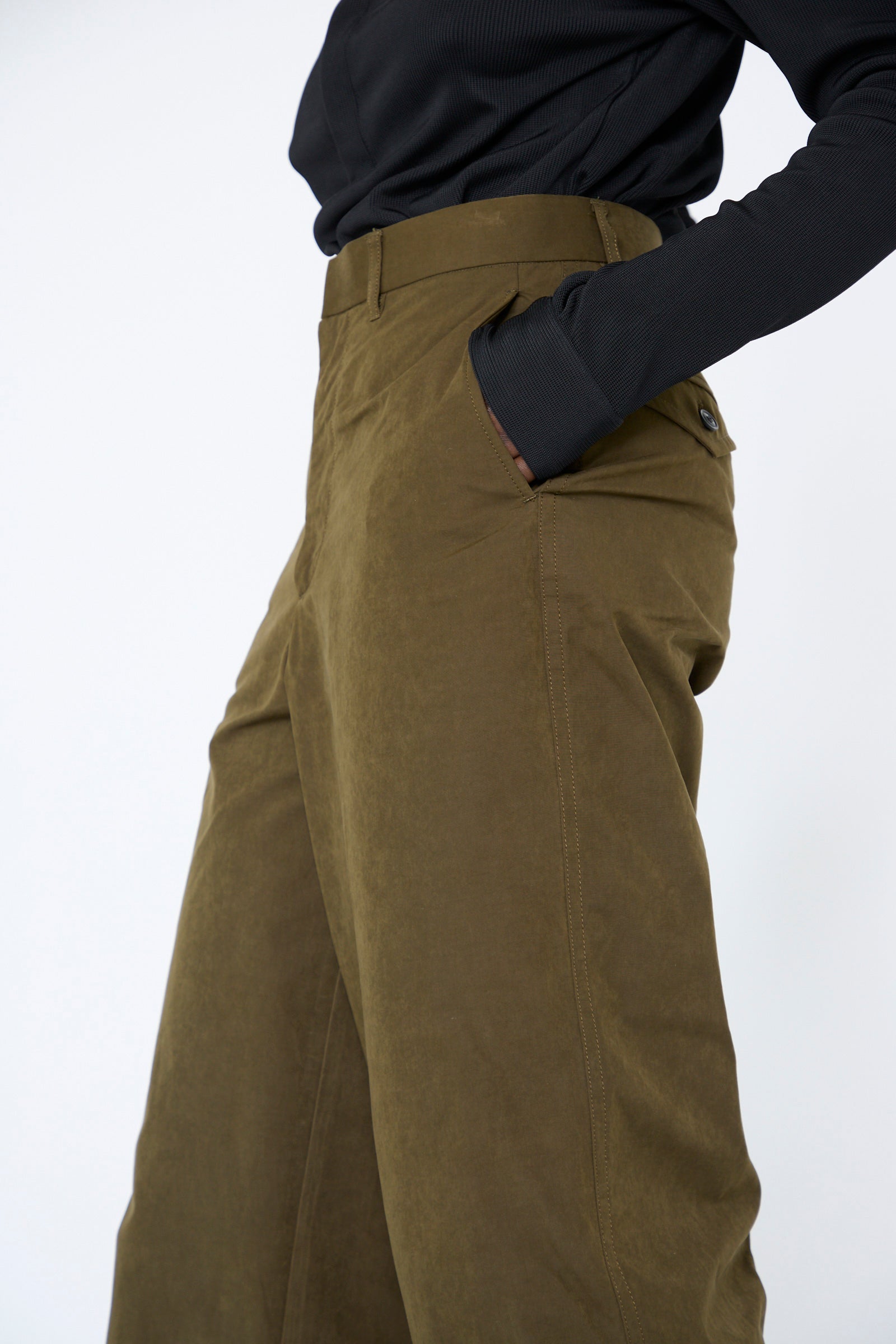 A person wearing a black long-sleeve top and TOGA ARCHIVES' Polyester Cotton Pant in Khaki, characterized by a high-waist, relaxed-fit, and wide-leg design, stands with one hand in their pocket.