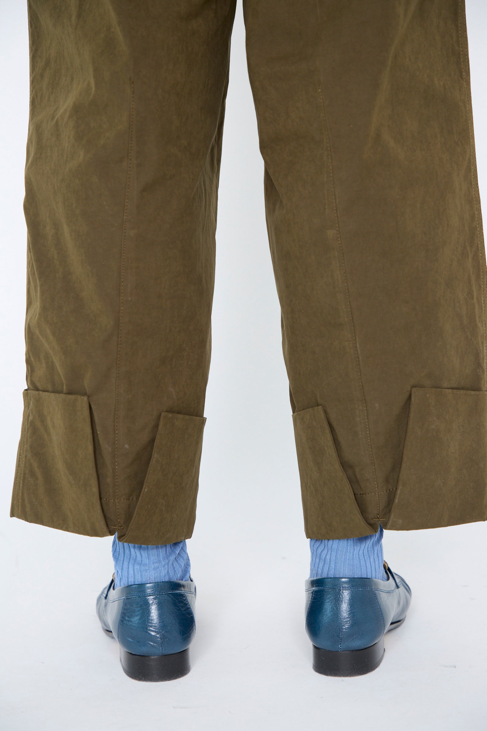 A person wearing TOGA ARCHIVES' Polyester Cotton Pant in Khaki, featuring a relaxed fit with cuffs and two back slits, stands on a light-colored surface. Paired  with blue socks and shiny blue shoes.