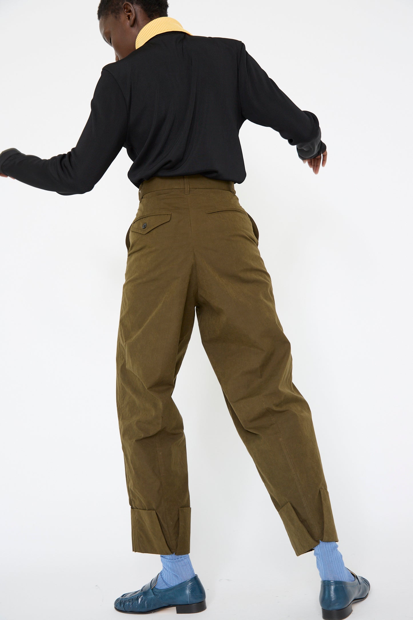 A person wearing a black shirt, relaxed fit Polyester Cotton Pants in khaki by TOGA ARCHIVES, blue socks, and blue shoes is facing away from the camera with one arm slightly bent.