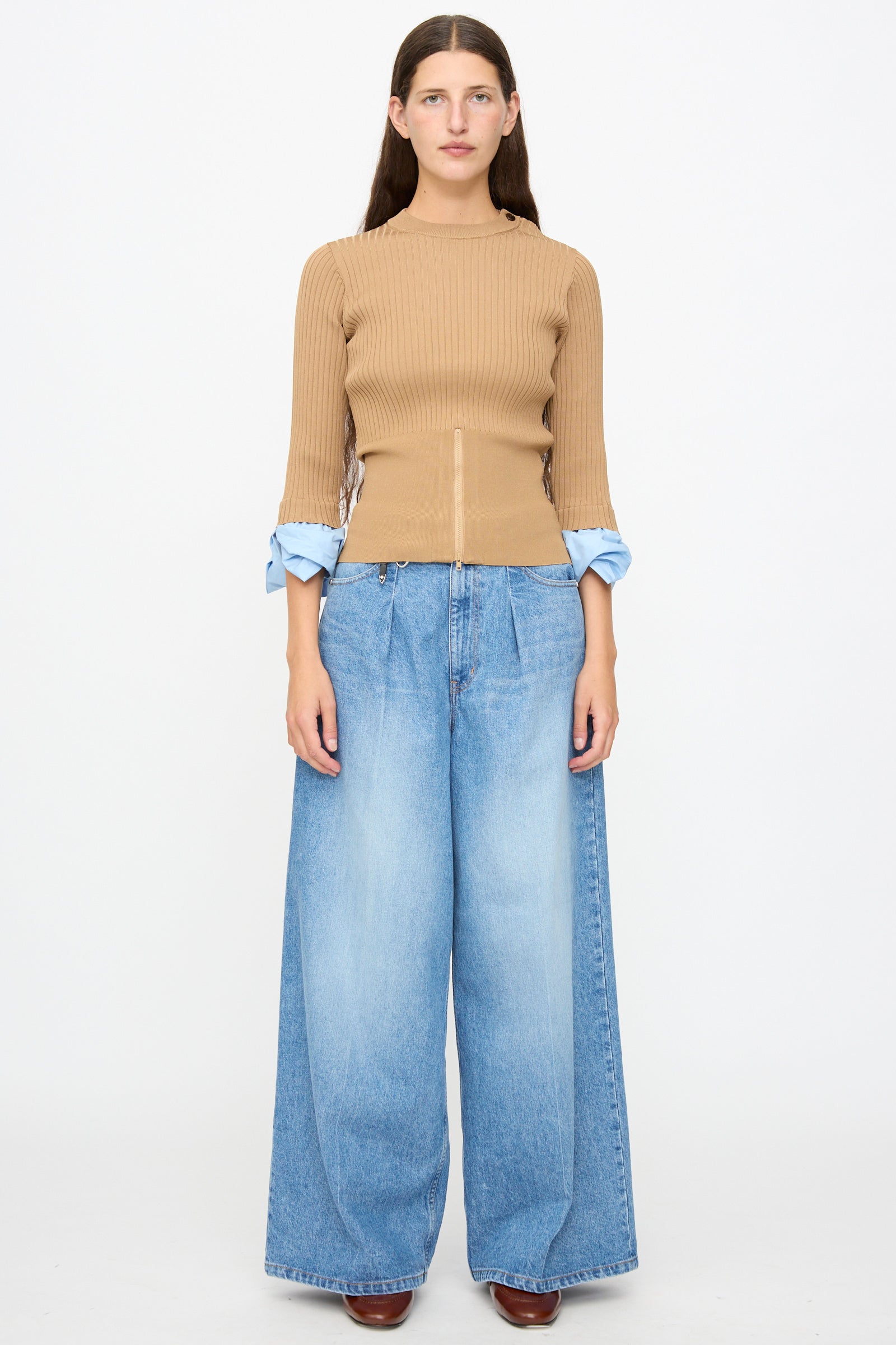 A person stands facing forward against a plain white background, wearing the TOGA ARCHIVES Rib Knit Top in Camel layered over a blue shirt and paired with wide-leg blue jeans.