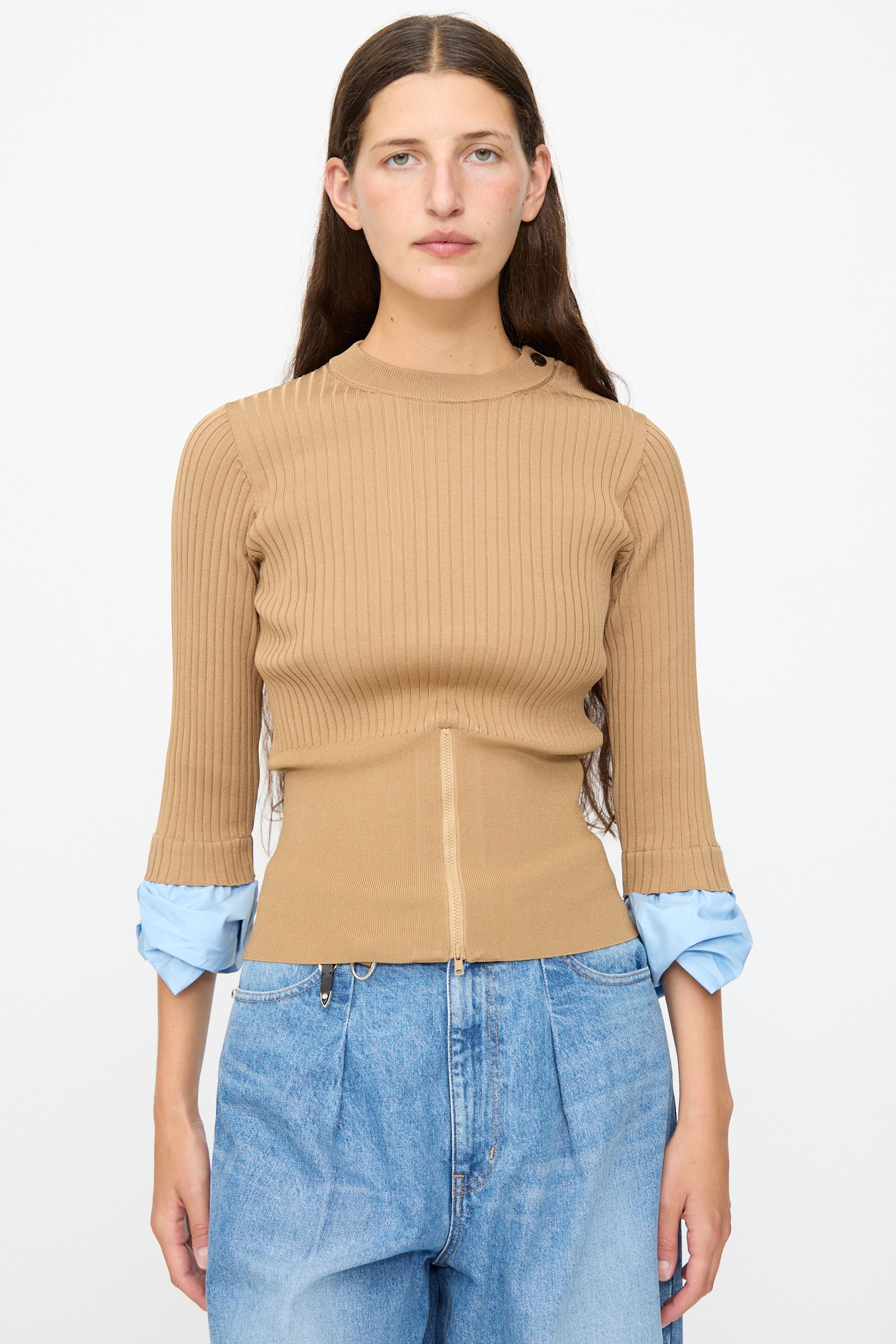 A woman with long brown hair wears the TOGA ARCHIVES Rib Knit Top in Camel, featuring blue cuffs, paired with light blue jeans and standing against a plain white background.