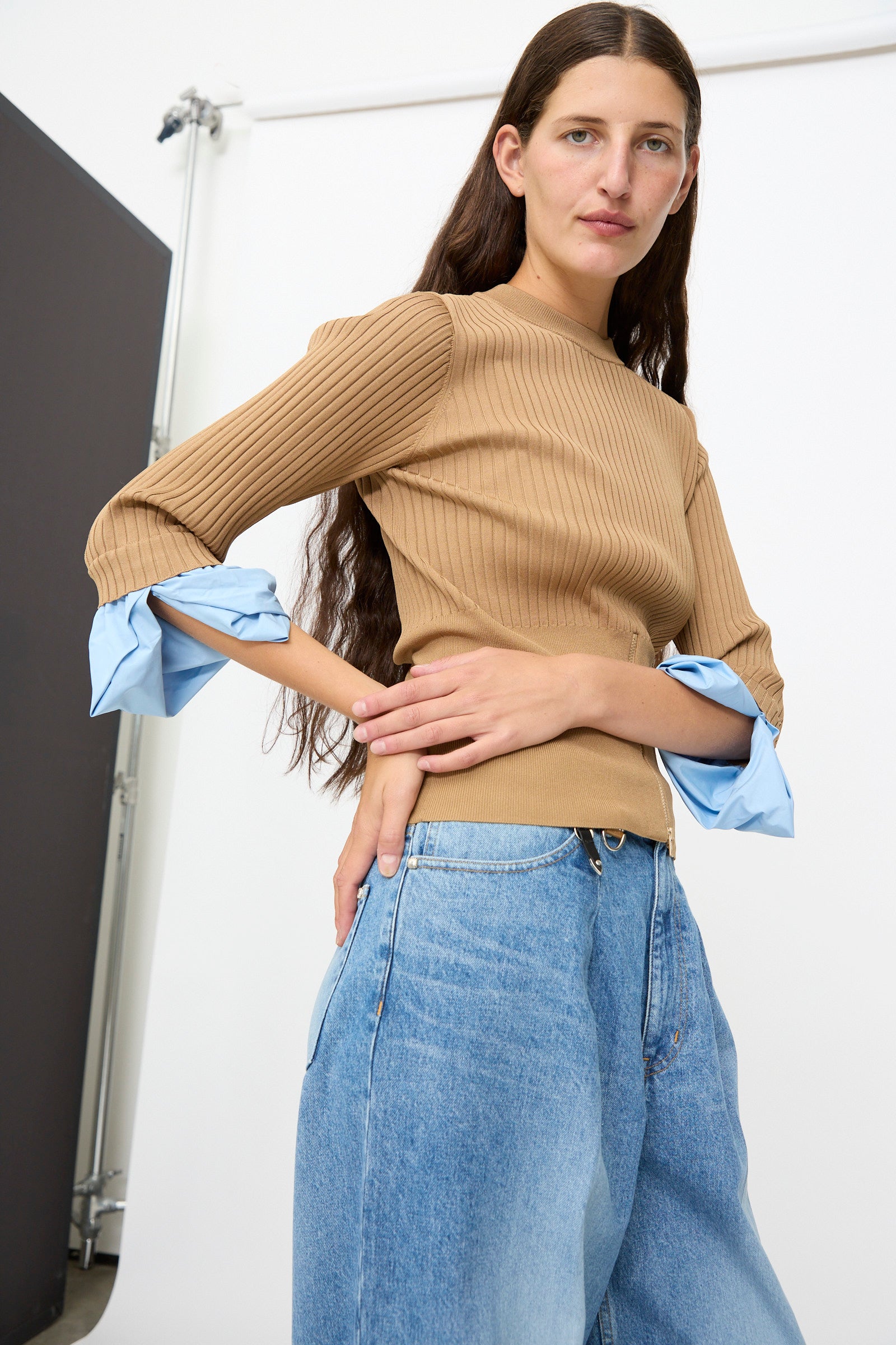 A woman with long brown hair wears the TOGA ARCHIVES Rib Knit Top in Camel—a slim fit sweater with blue cuffs—paired with high-waisted blue jeans, standing against a white and black studio background.