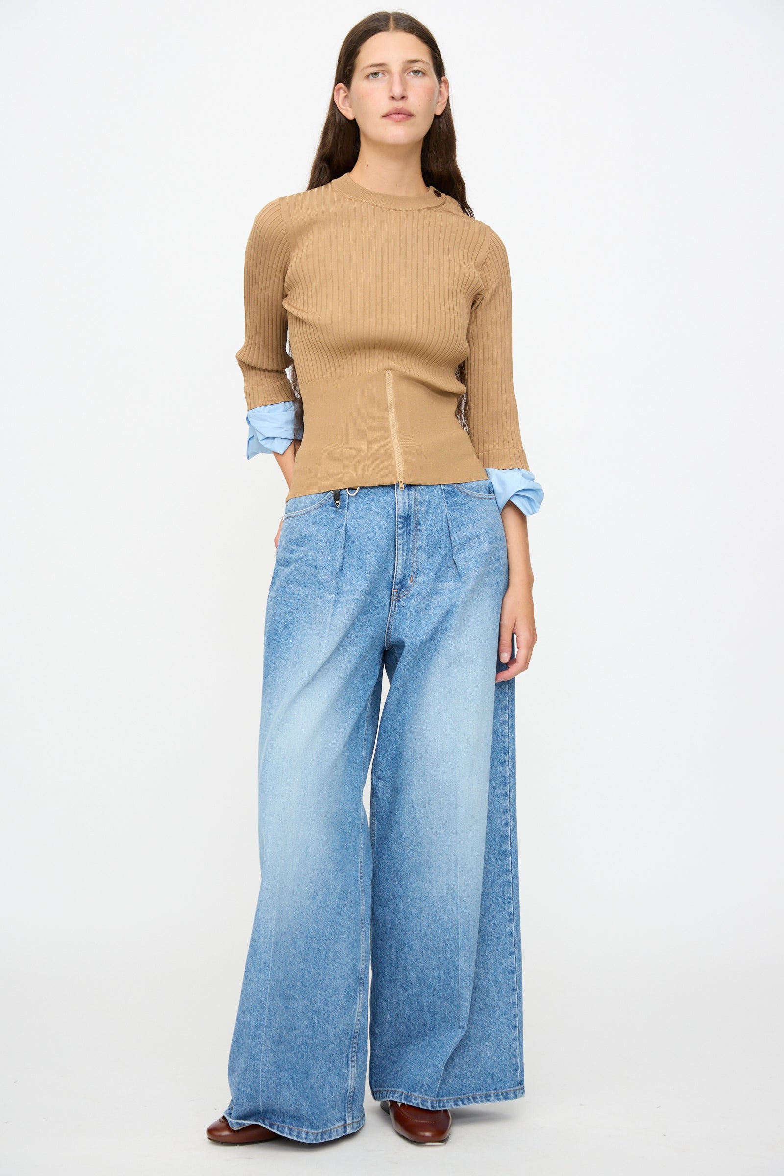 A person stands against a plain background, wearing the TOGA ARCHIVES Rib Knit Top in Camel layered over a blue shirt, paired with wide-leg blue jeans and brown shoes.