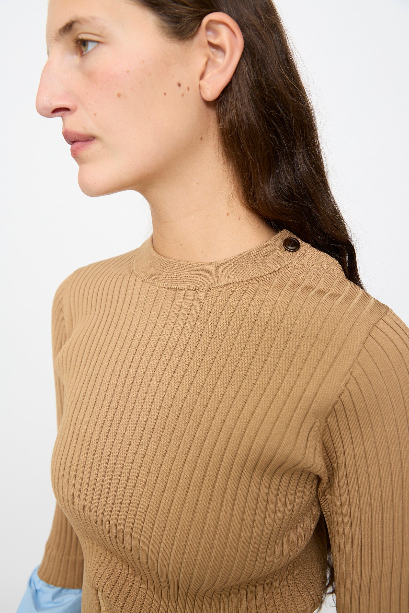 A person with long brown hair, shown from the side against a plain background, is wearing the TOGA ARCHIVES Rib Knit Top in Camel—a slim-fit, tan long-sleeve pullover sweater featuring rib knit texture and button detailing on the shoulder.
