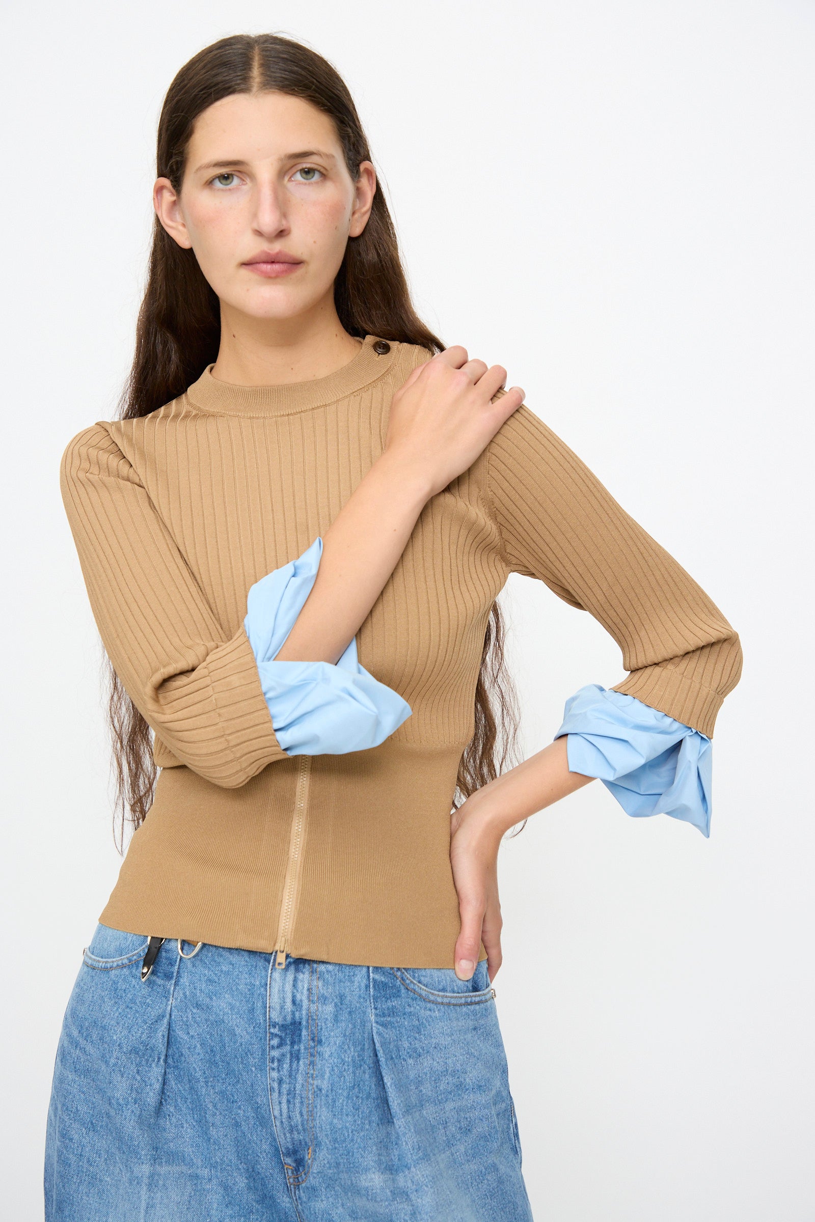 A woman with long brown hair is wearing the TOGA ARCHIVES Rib Knit Top in Camel, featuring blue cuffs, paired with light blue jeans, and standing against a plain white background.