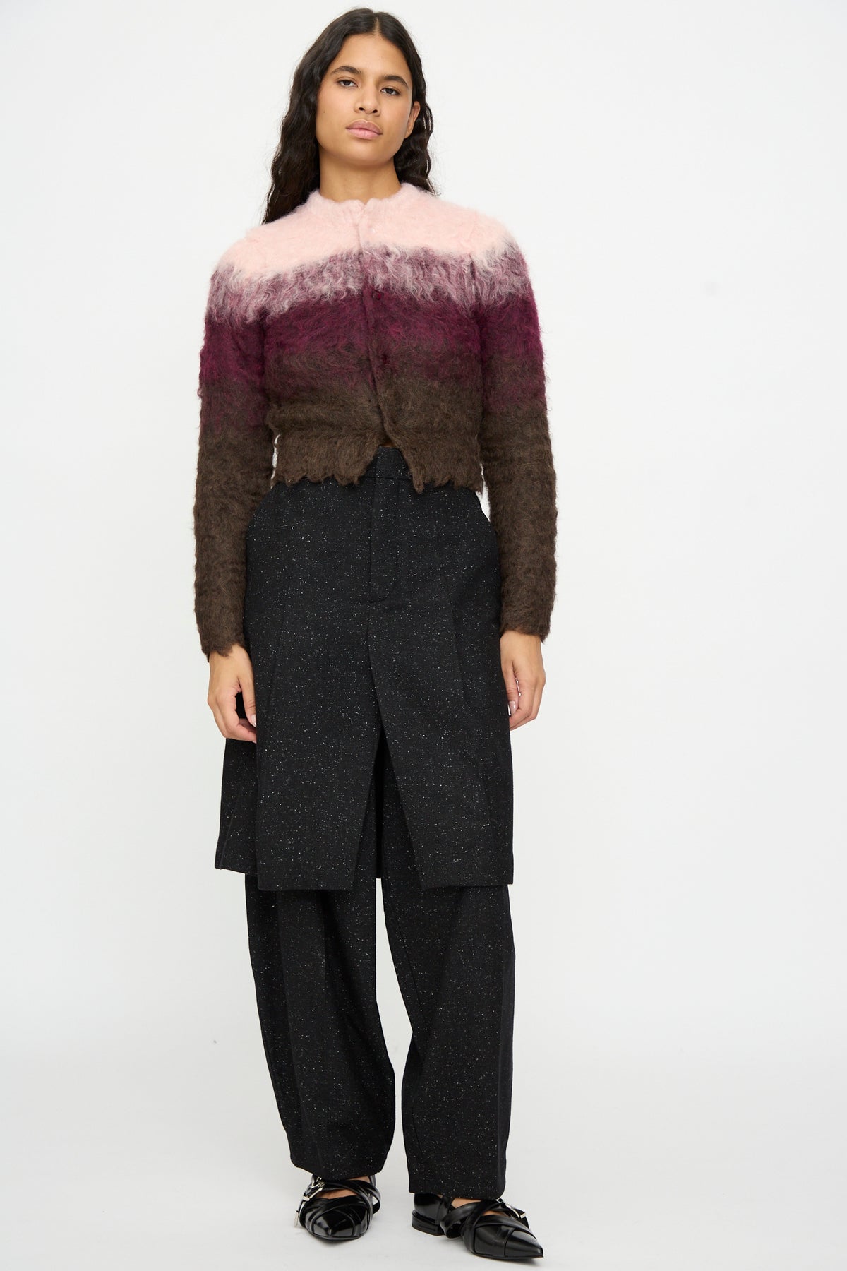 A person stands against a white background, wearing the TOGA ARCHIVES Shaggy Knit Cardigan in Pink paired with black layered pants and black shoes.