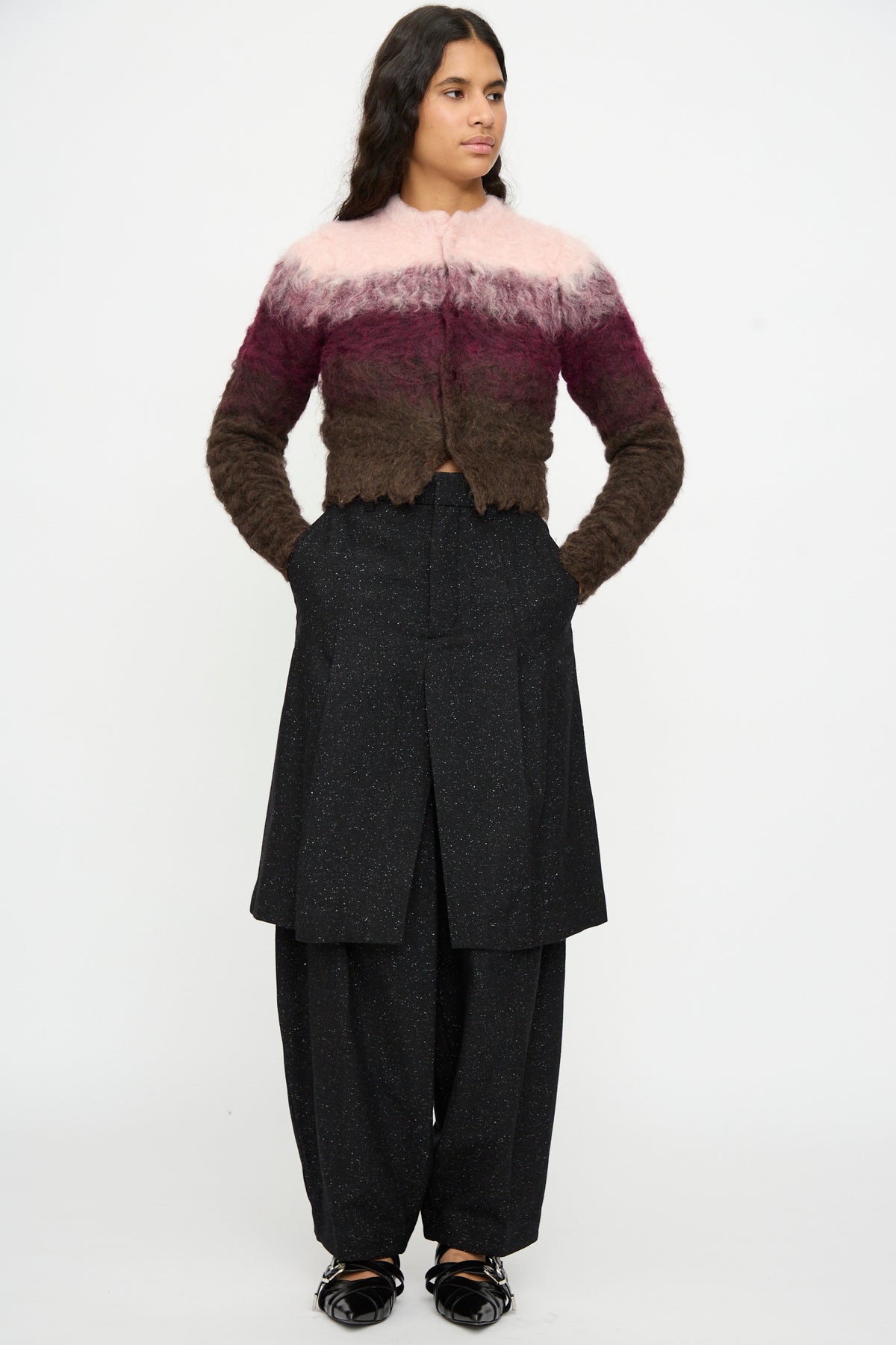 A person stands against a plain background wearing the TOGA ARCHIVES Shaggy Knit Cardigan in Pink, paired with wide-leg black pants featuring layered panels and black shoes. Their hands are in their pockets.