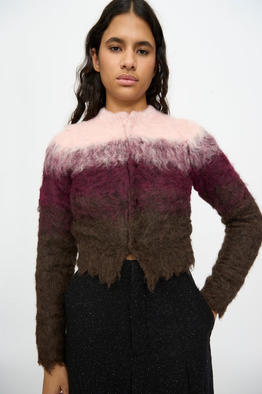 A person wearing the TOGA ARCHIVES Shaggy Knit Cardigan in Pink—featuring pink, maroon, and brown color gradients—paired with black pants, stands against a plain white background.