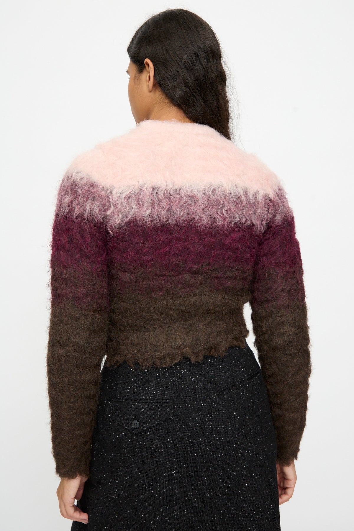 A woman stands facing away, wearing the TOGA ARCHIVES Shaggy Knit Cardigan in Pink—featuring pink, burgundy, and brown horizontal stripes—paired with a black skirt.