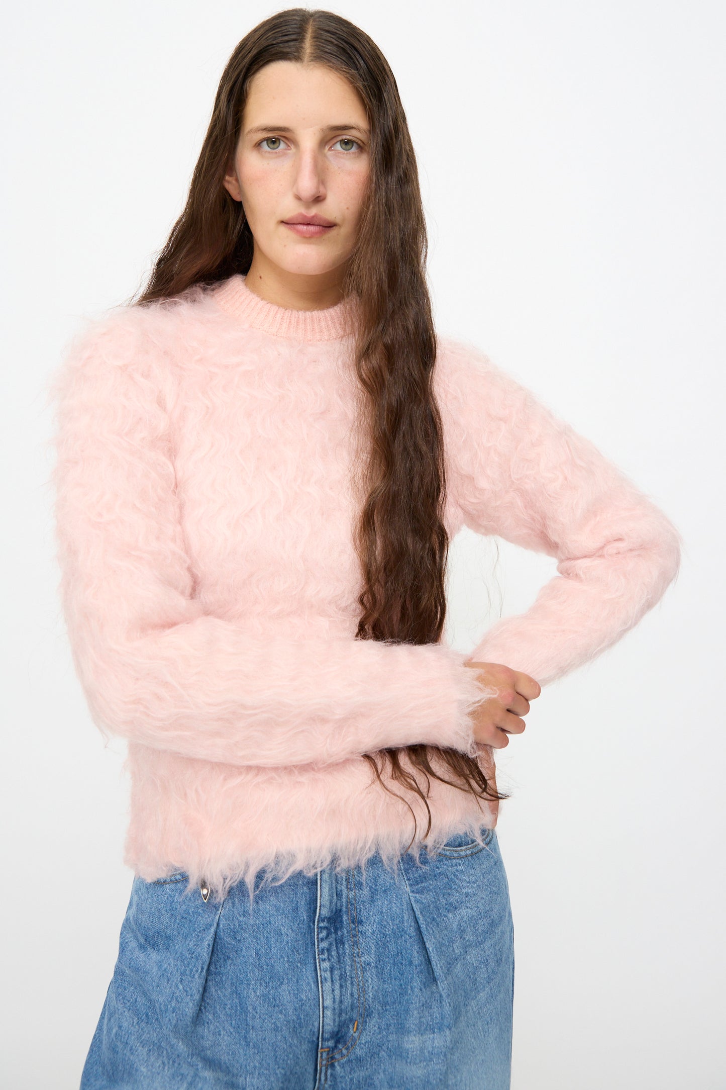 A person with long brown hair is wearing the TOGA ARCHIVES Shaggy Knit Pullover in Pink styled in a relaxed fit, paired with blue jeans and standing against a plain white background.
