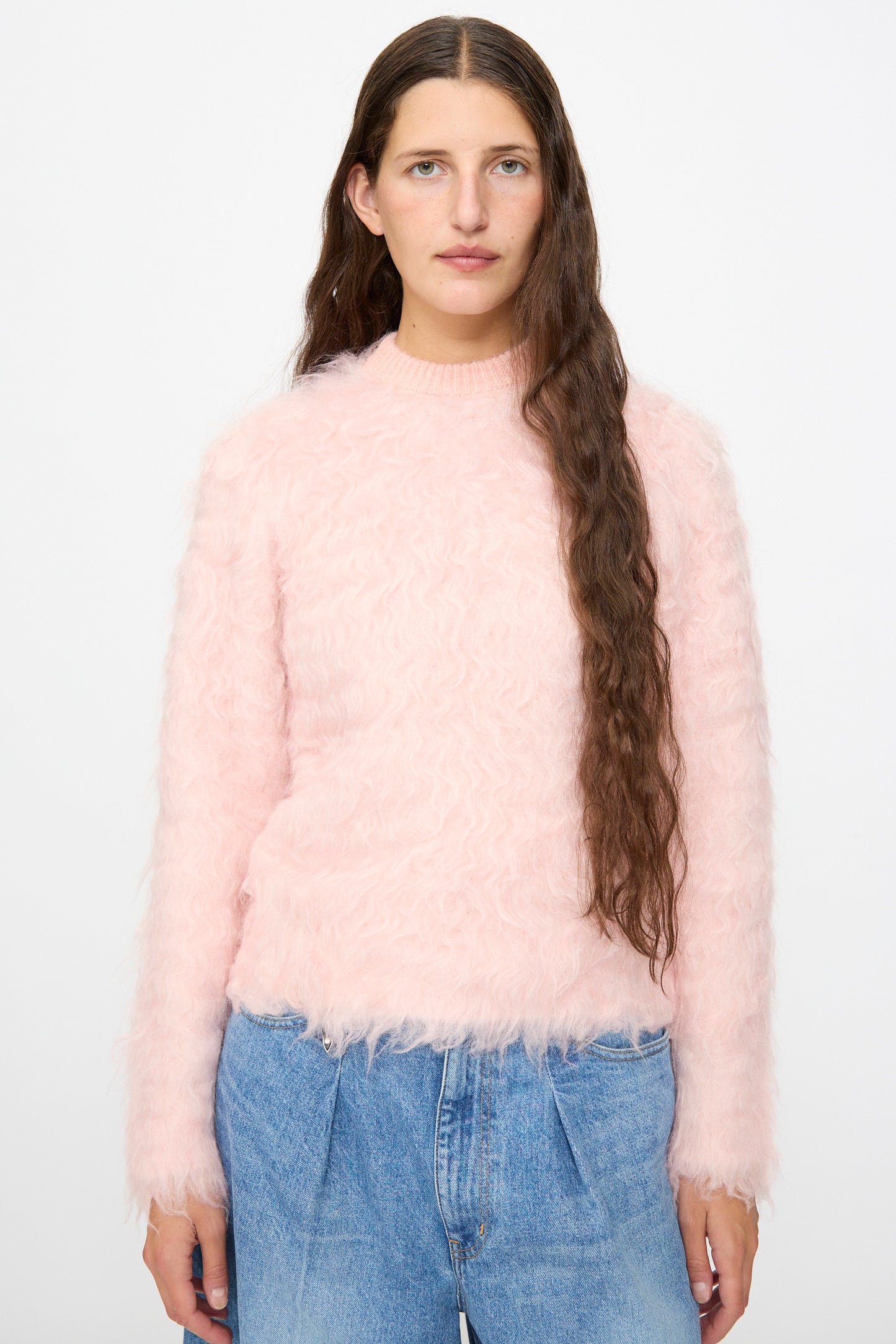 A person with long brown hair wears the TOGA ARCHIVES Shaggy Knit Pullover in Pink, paired with blue jeans, standing against a plain white background.