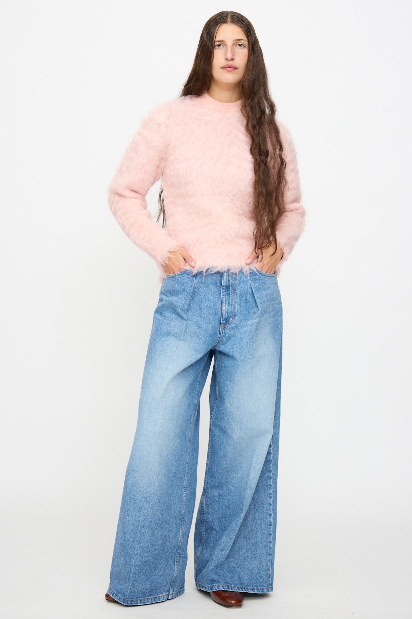 A person stands against a white background wearing the TOGA ARCHIVES Shaggy Knit Pullover in Pink and wide-leg blue jeans, with their hands in their pockets.