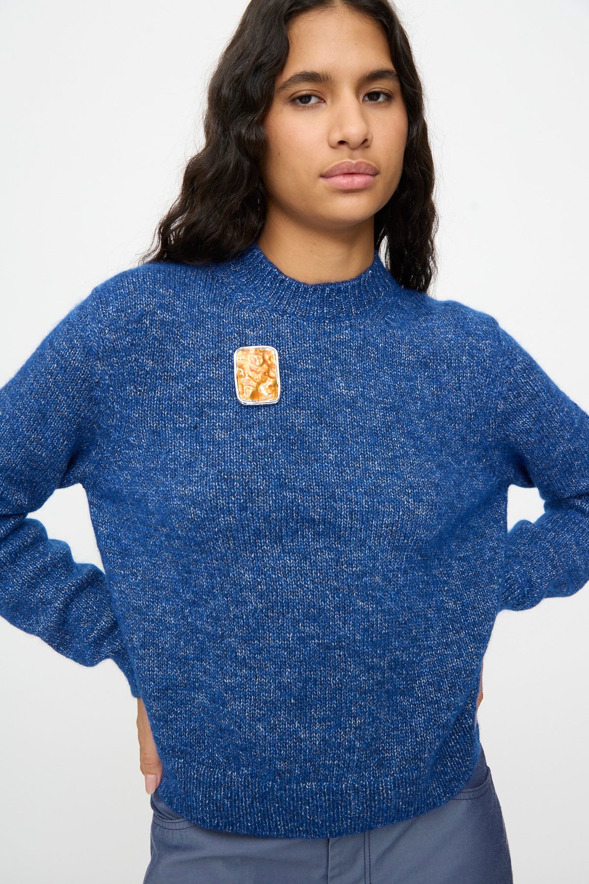 A person wearing a blue knit sweater with hands on hips stands against a plain background, styled with the TOGA ARCHIVES Square Wave Brooch in Orange. The striking brooch features an elegant orange accent complemented by silver-tone brass details, adding a refined touch to the overall look.