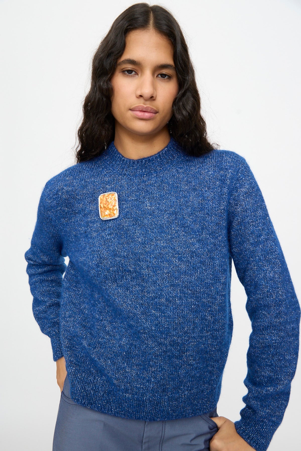 A person with long dark hair wearing a blue sweater is shown standing against a plain light background, with the TOGA ARCHIVES Square Wave Brooch in Orange—crafted in Japan—pinned on the left side.