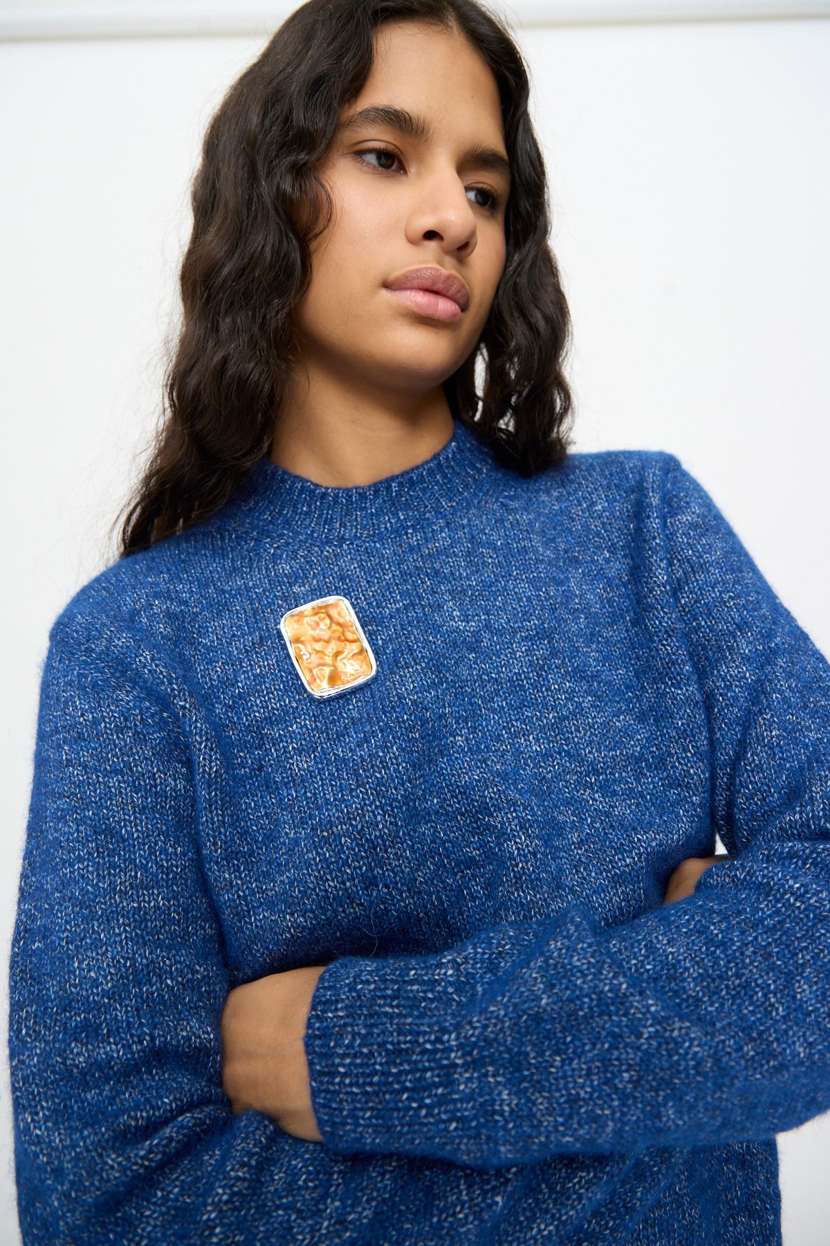 A person with long wavy hair, dressed in a blue sweater and wearing the TOGA ARCHIVES Square Wave Brooch in Orange, stands with arms crossed against a plain background.