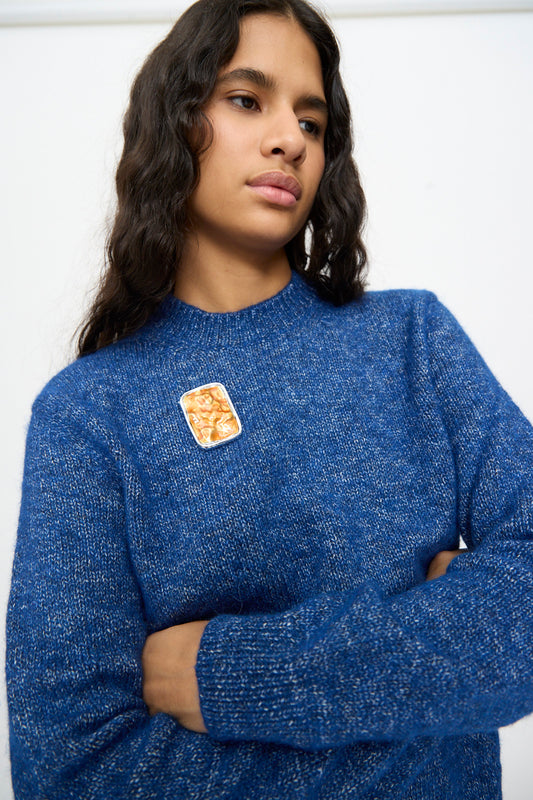A person with long wavy hair, dressed in a blue sweater and wearing the TOGA ARCHIVES Square Wave Brooch in Orange, stands with arms crossed against a plain background.