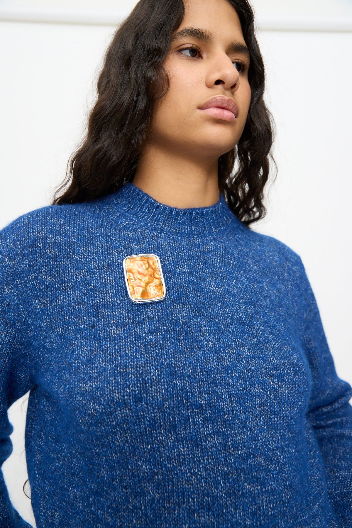 A person wearing a textured blue sweater with a TOGA ARCHIVES Square Wave Brooch in Orange—a rectangular, silver-tone brass brooch featuring an abstract design—on the chest, made in Japan.