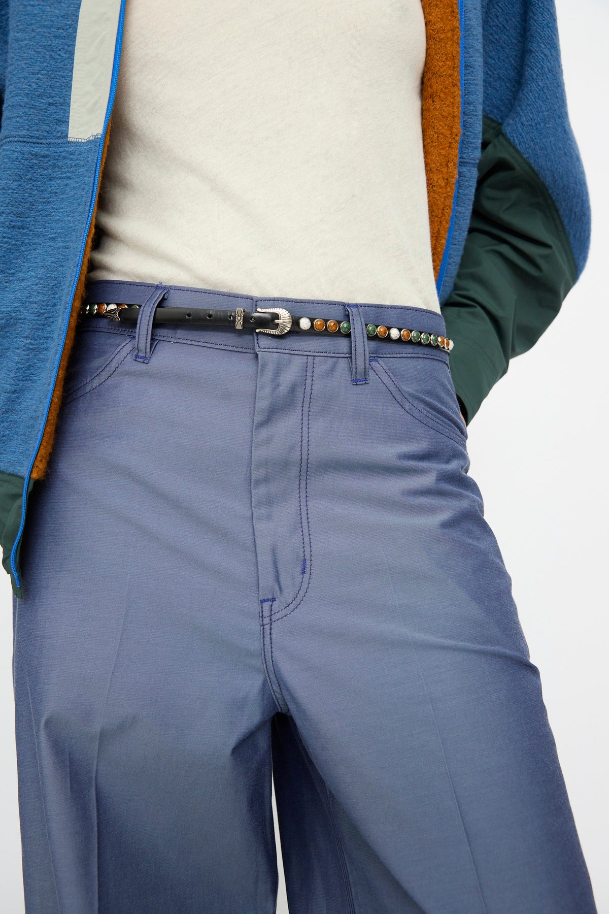 A person is shown from the waist to mid-thigh wearing a cream shirt, blue jacket, and blue pants, styled with the TOGA ARCHIVES Stone Leather Belt in Black—an adjustable fit leather belt adorned with multicolored beads and made in Japan.