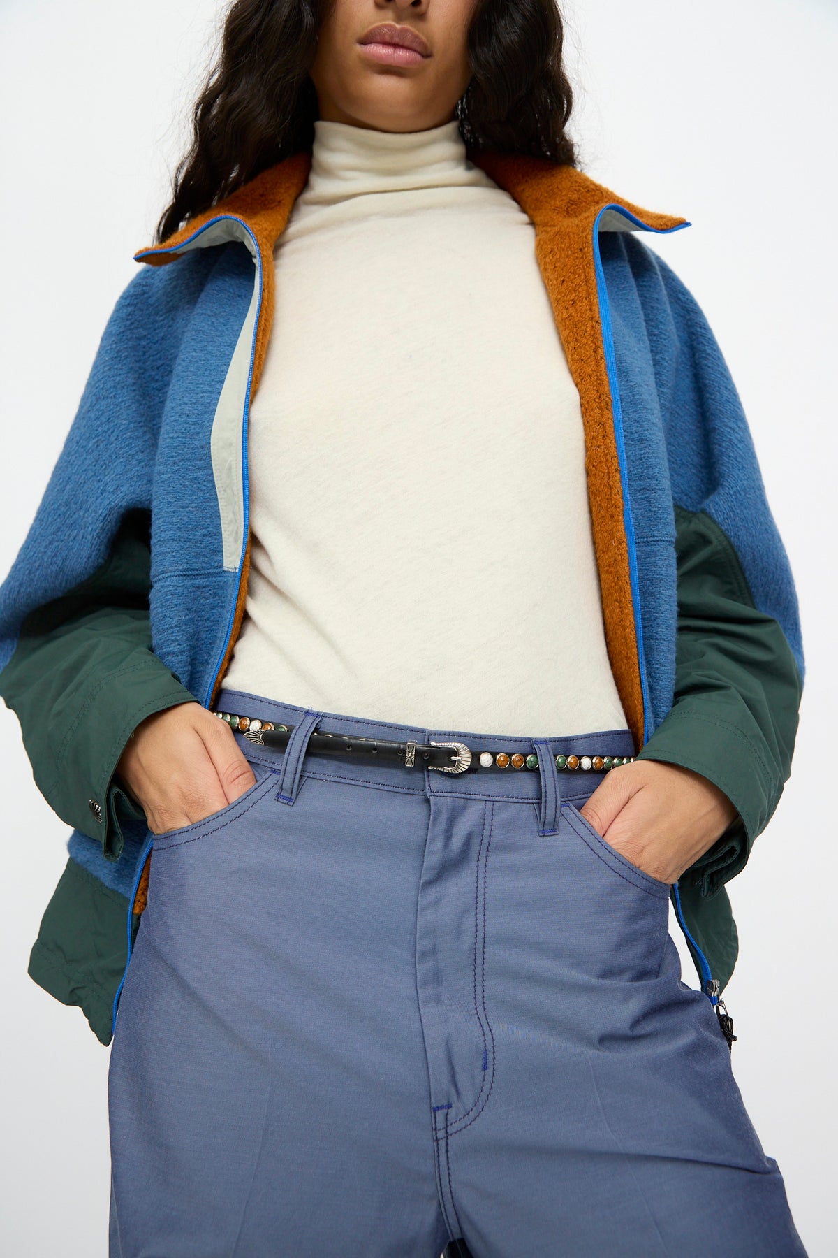 Outfitted in a cream turtleneck, blue and green color-block jacket, blue pants, and the TOGA ARCHIVES Stone Leather Belt in Black—featuring metallic accents for an adjustable fit—the person stands with hands in their pockets.