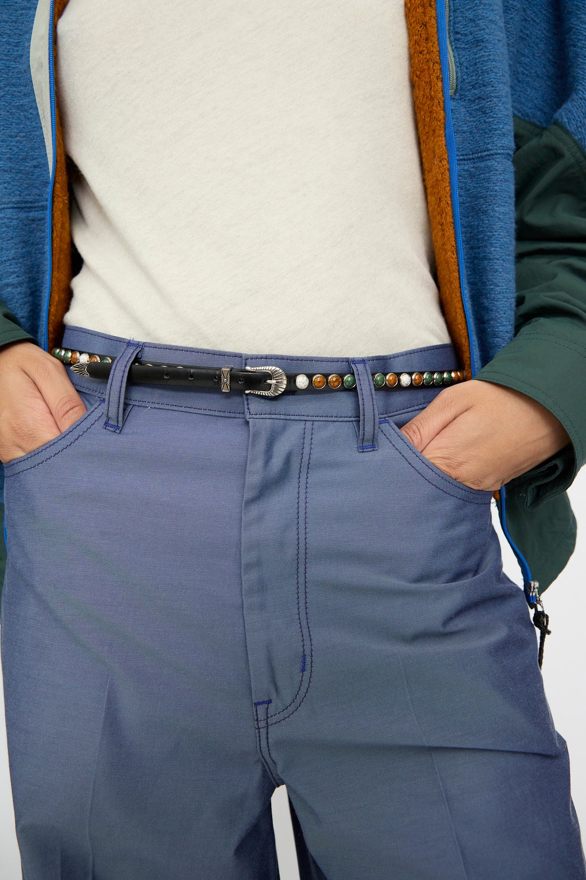 A person wears blue pants, a white shirt, a blue and green jacket, and the TOGA ARCHIVES Stone Leather Belt in Black—a black leather belt featuring decorative studs and an adjustable fit—with their hands in their pockets.
