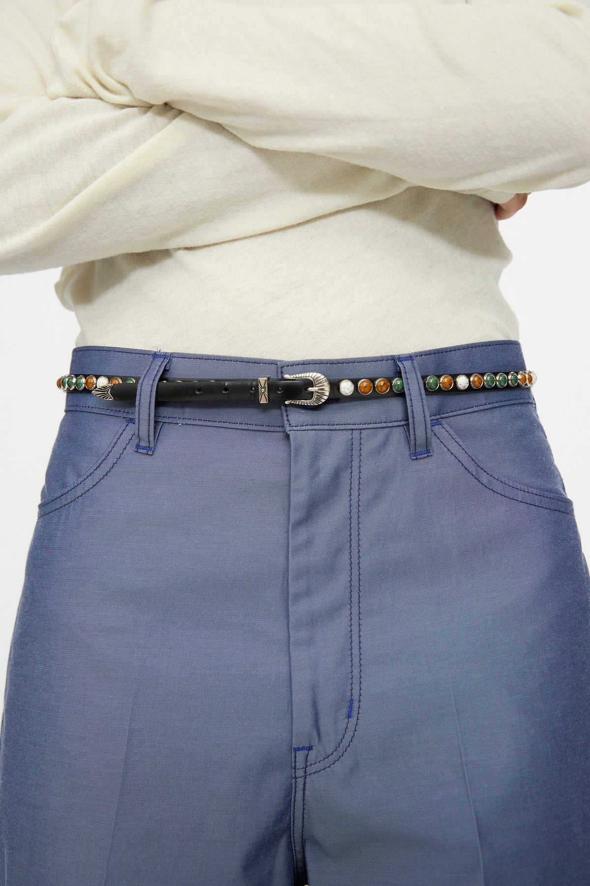 A person with arms crossed wears a cream long-sleeve top, blue pants, and the TOGA ARCHIVES Stone Leather Belt in Black, featuring colorful round studs and an adjustable fit.
