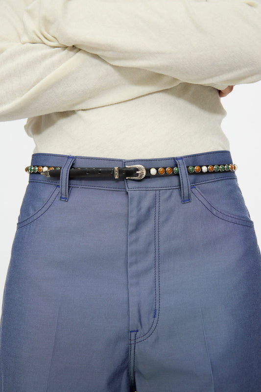 A person with arms crossed wears a cream long-sleeve top, blue pants, and the TOGA ARCHIVES Stone Leather Belt in Black, featuring colorful round studs and an adjustable fit.