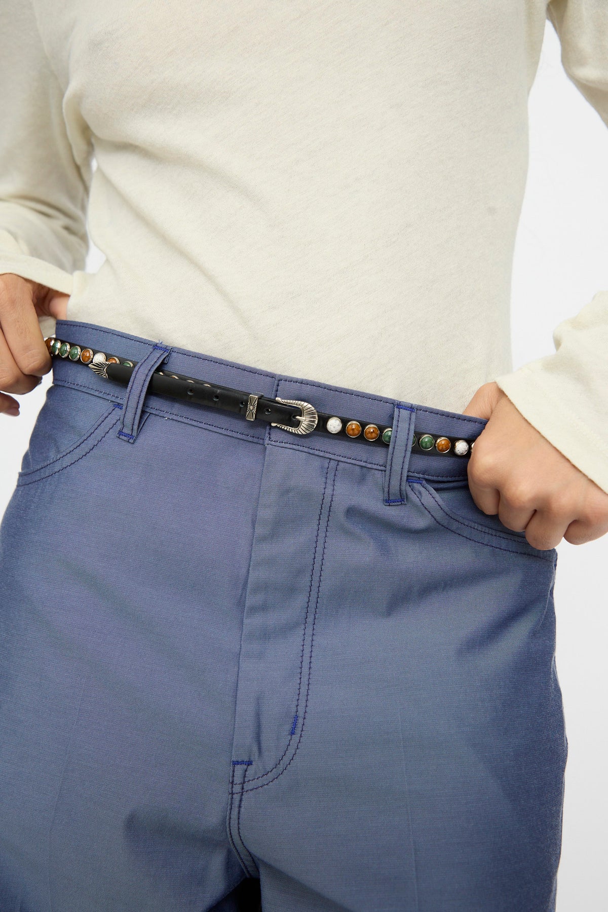 Wearing a light long-sleeve shirt and blue pants, the person adjusts the TOGA ARCHIVES Stone Leather Belt in Black—an adjustable-fit black leather belt adorned with colorful round studs, made in Japan.